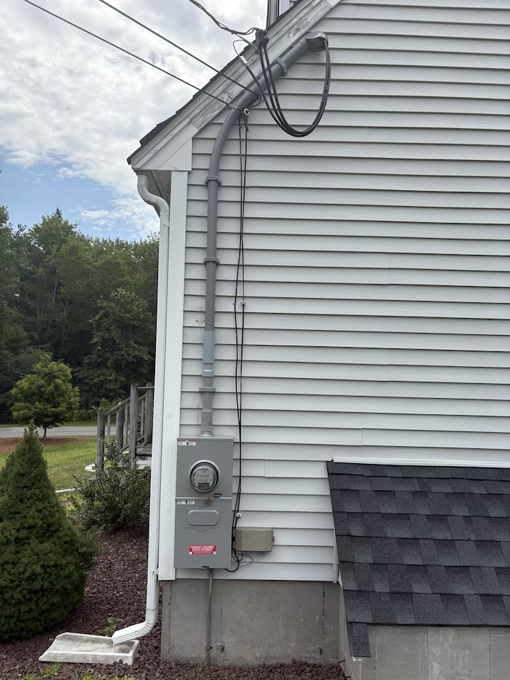 Close-up of the side of a house with gray vinyl siding, an electrical meter, and electrical wiring. There are trees and a cloudy sky in the background.