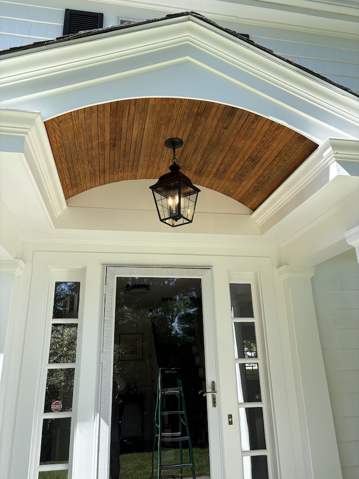 Close-up of the front porch of a house, featuring a wooden ceiling with a hanging lantern, white framing, glass door, and surrounding windows.