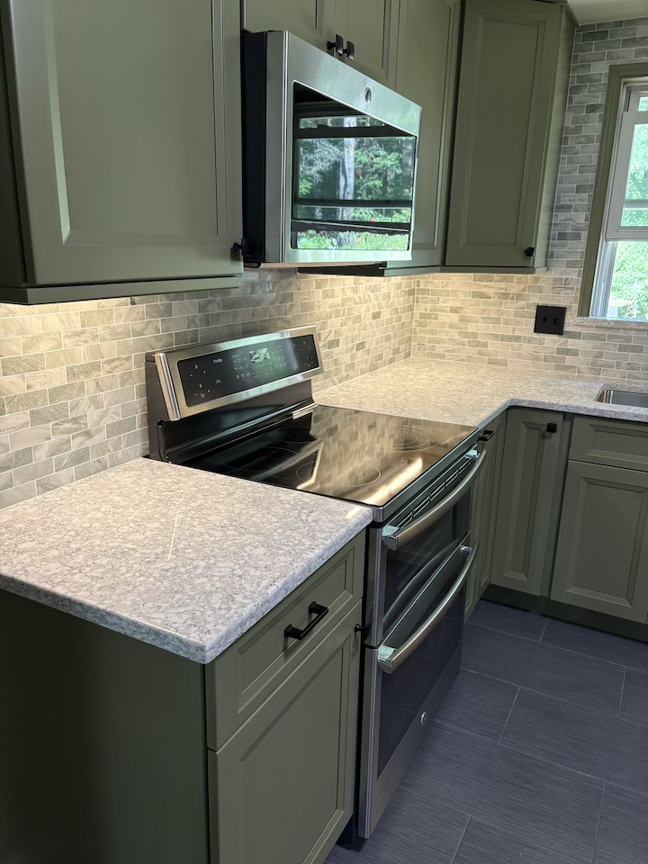 Kitchen with green cabinets, stainless steel oven and microwave, granite countertops, beige brick backsplash, and a window with trees outside.