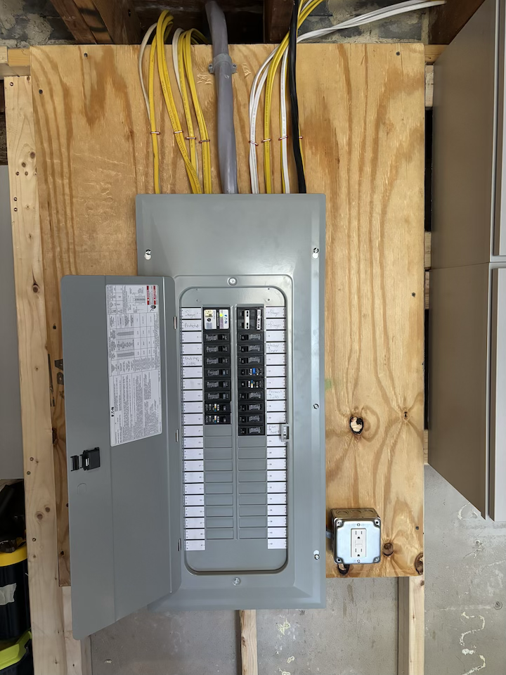 Electric breaker panel with yellow, white, gray, and black wires connected, mounted on a plywood wall in a basement or utility room.