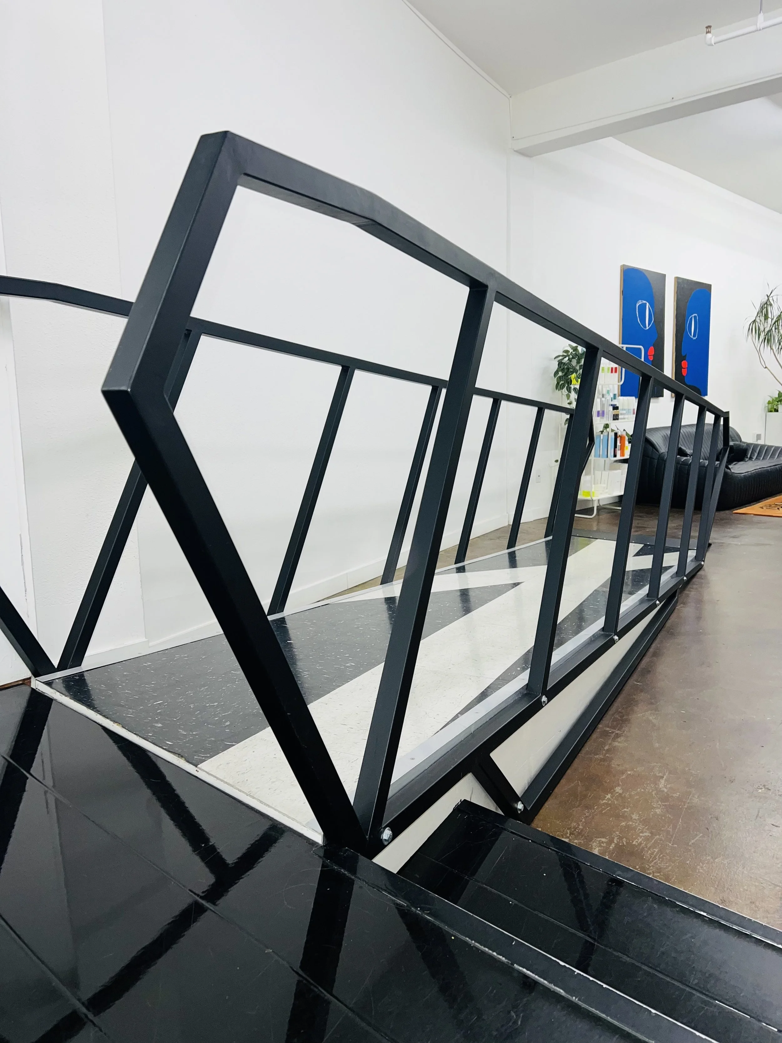 Black metal railing with a similar shape to a rhombus, separating a textured tile flooring area from a polished black tile floor, with a more decorated seating area and artwork in the background.