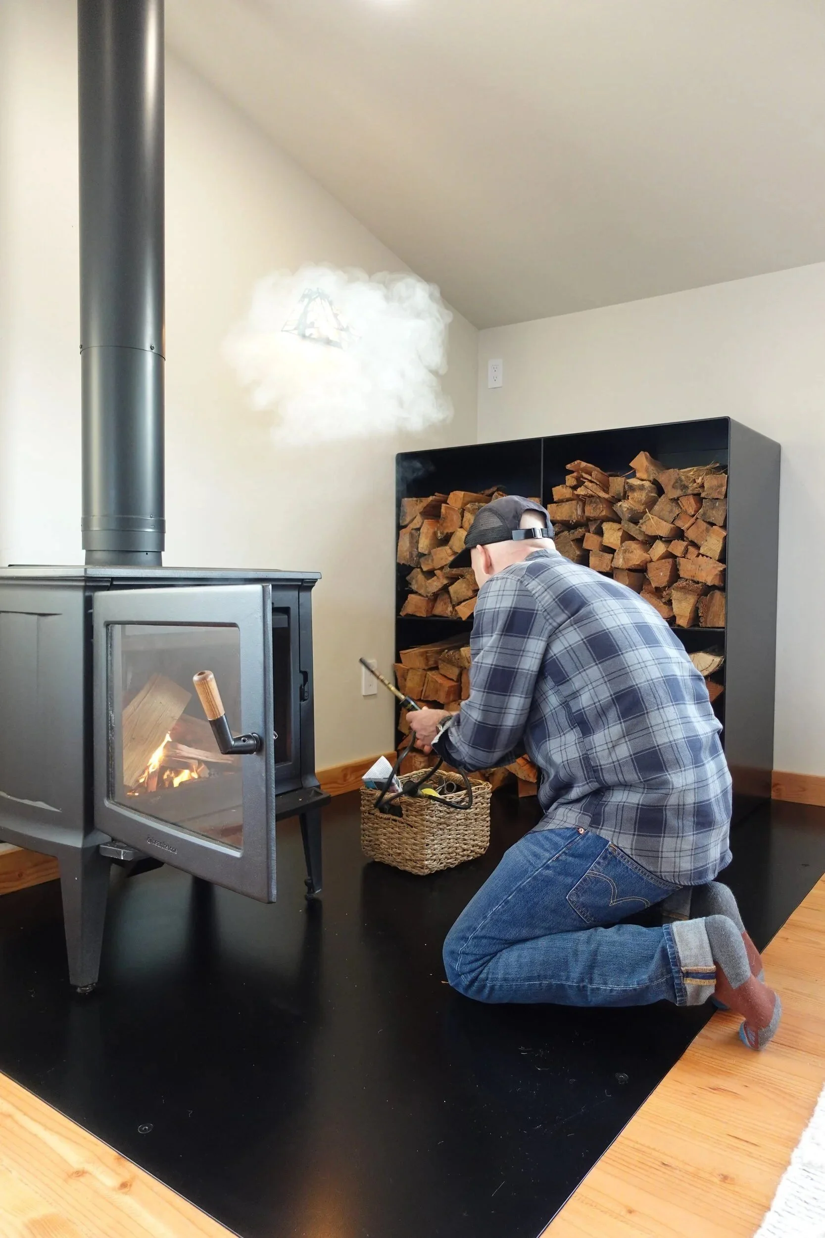 Homeowner lighting a wood stove beside a custom steel firewood box and flush steel hearth — designed and fabricated by de stijl FORMS in Hood River, Oregon.