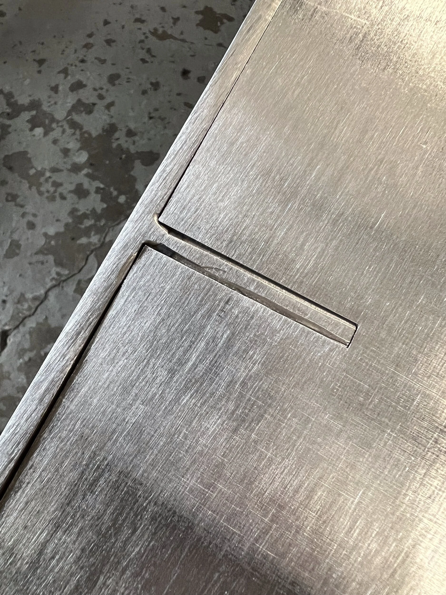 Back panel of a steel firewood box with a laser-cut tab locking into a slot in the top panel, a tab-and-slot joint plug welded and ground flush — designed and fabricated by de stijl FORMS in Hood River, Oregon.