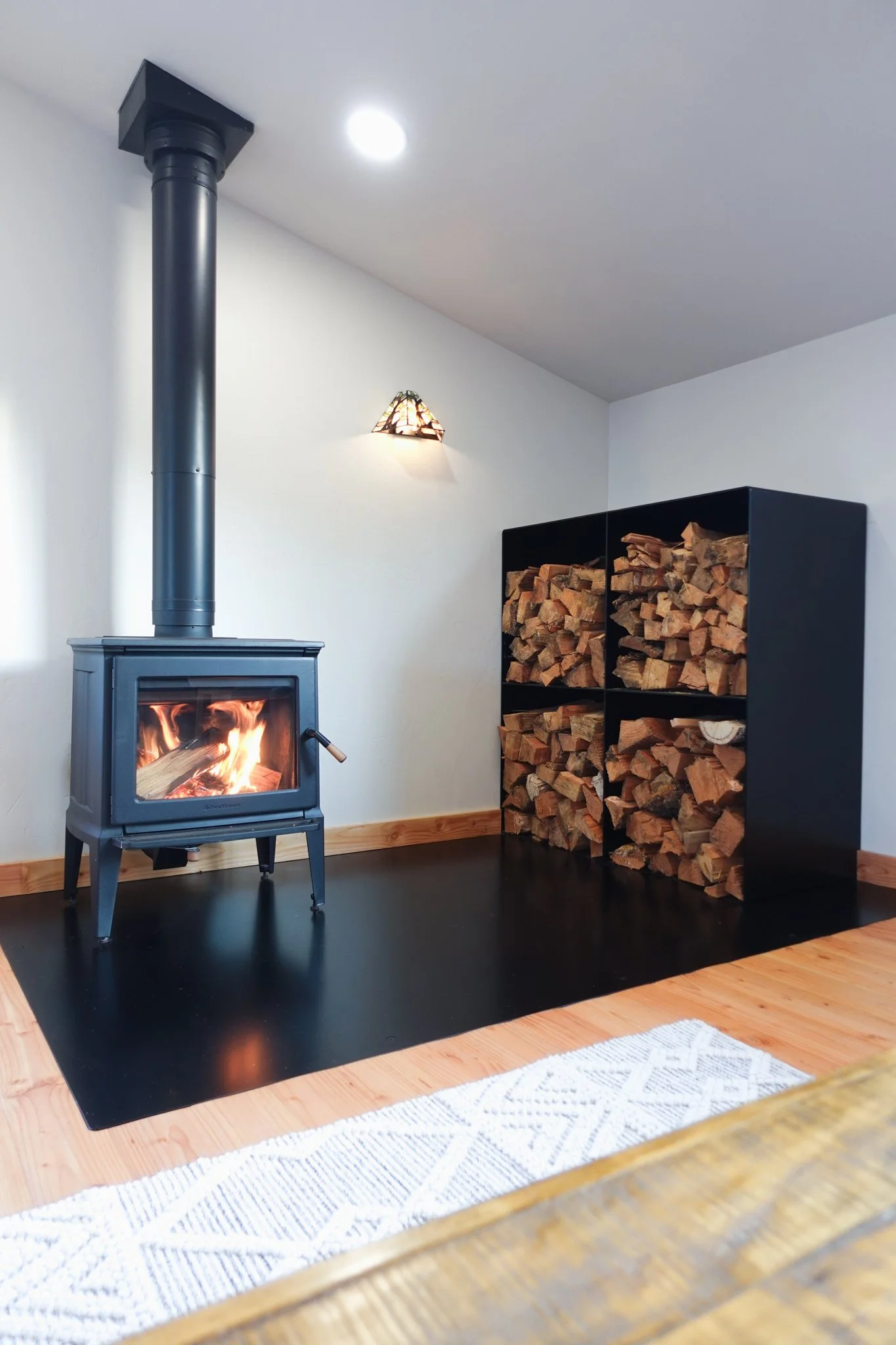 A cozy living room corner featuring a wood-burning stove with a fire burning inside, a black wood storage cube filled with stacked firewood, a small wall-mounted lamp, and a white rug on a wooden floor.