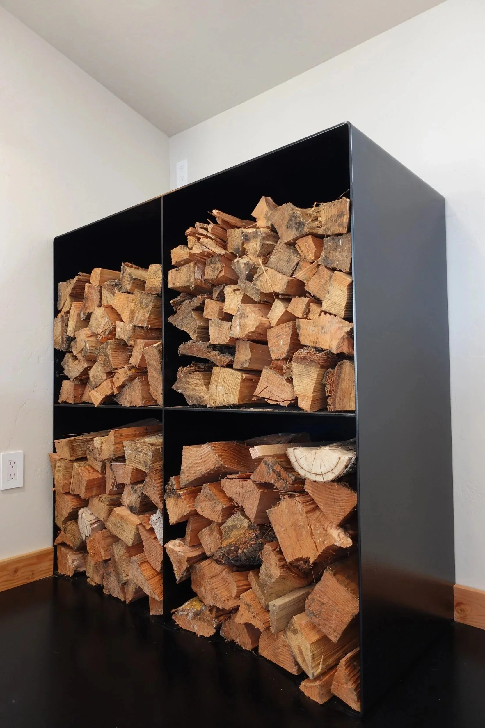 Straight-on view of a satin black steel firewood box stocked with split wood — designed and fabricated by de stijl FORMS in Hood River, Oregon.