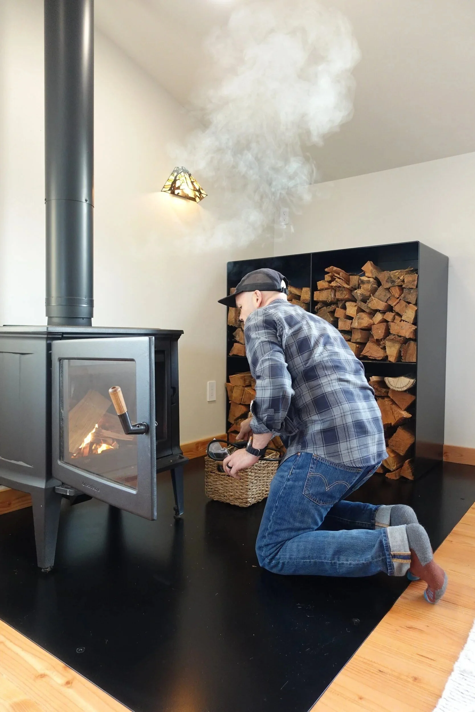 Lit wood stove with a fire burning next to a stocked satin black steel firewood box — designed and fabricated by de stijl FORMS in Hood River, Oregon.