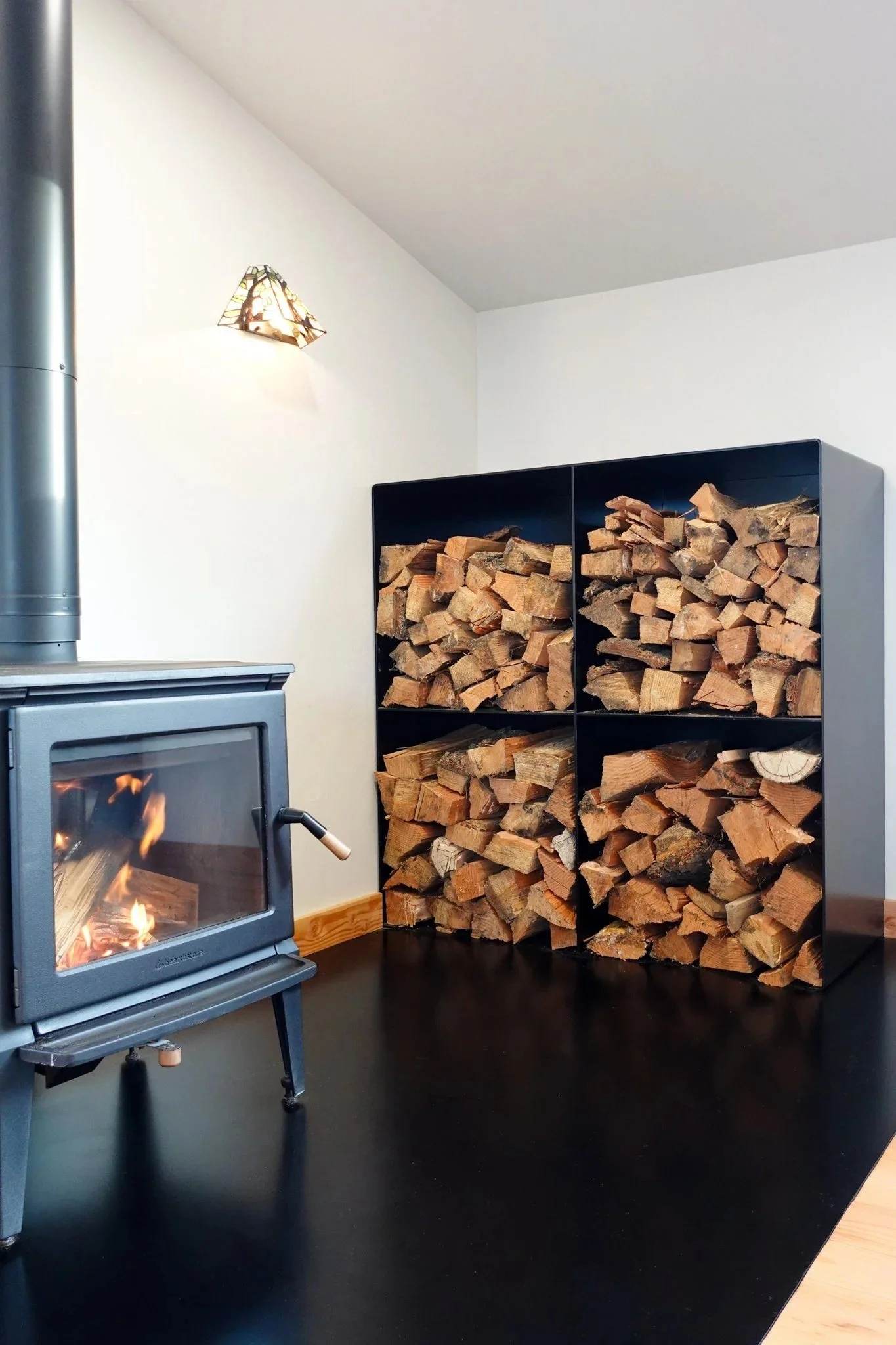 Custom steel firewood storage box with folded steel shelves, stocked with split firewood next to a wood stove — designed and fabricated by de stijl FORMS in Hood River, Oregon.