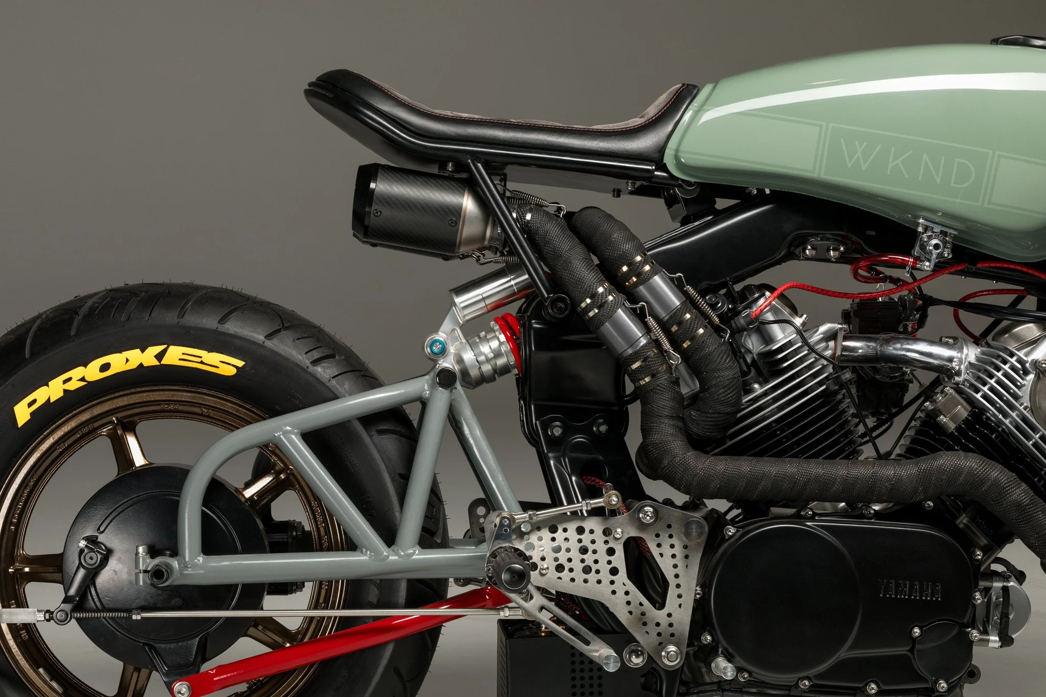 Close-up of a custom motorcycle showing the rear wheel, seat, and engine components, with a grey frame and black, silver, and red accents.