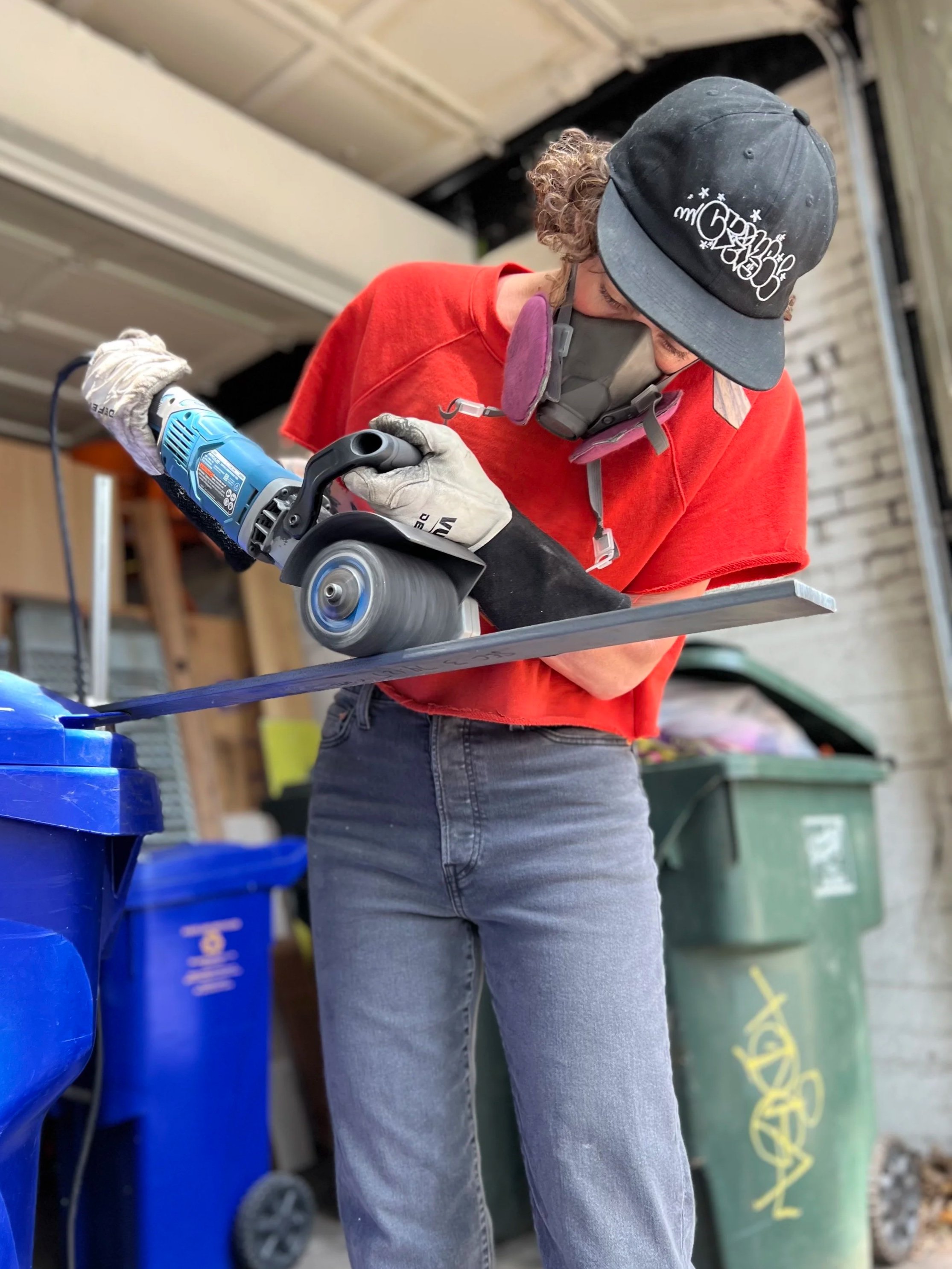 Person wearing a black cap, gray mask, and gloves, using a handheld power tool to cut a piece of metal or wood in a workshop or garage.