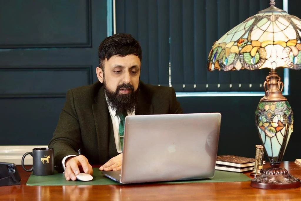 A man with dark hair and a beard, wearing a black blazer, is sitting at a wooden desk working on a silver laptop. The desk has a black coffee mug with a gold dog emblem, a green mouse pad, a computer mouse, some books, and a decorative Tiffany-style lamp with colorful stained glass. The background has dark blue paneling and window blinds.