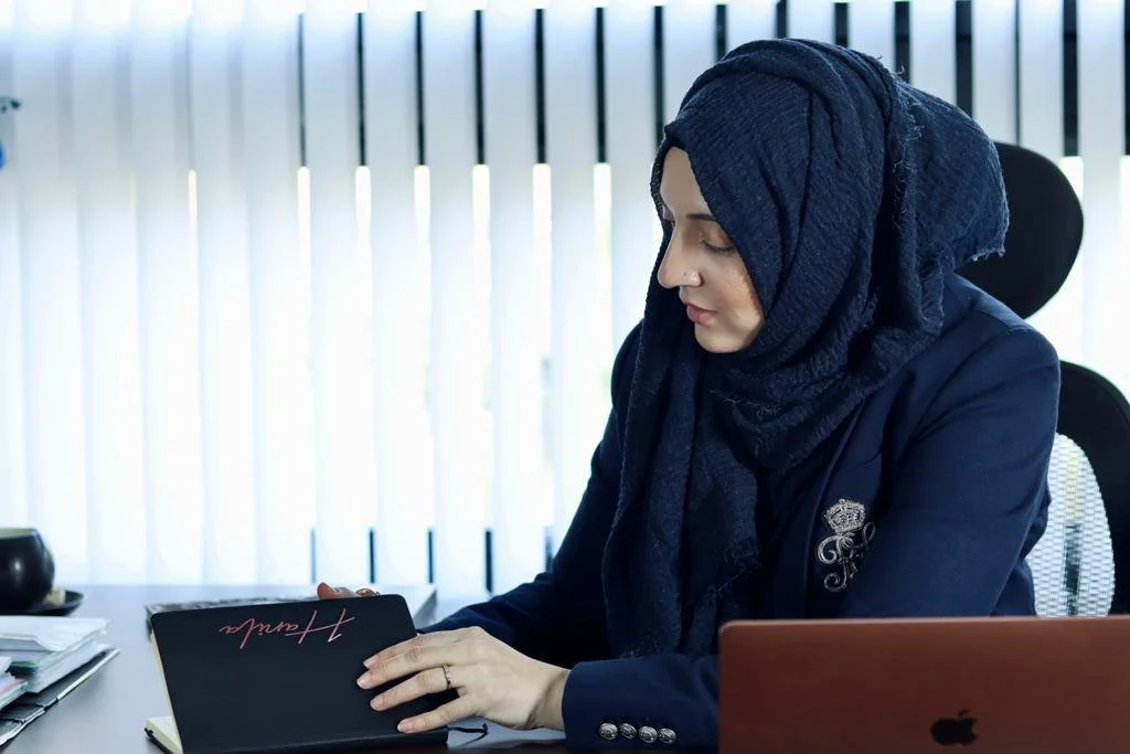 A woman wearing a navy hijab and navy blazer sitting at a desk indoors, looking at a black notebook.