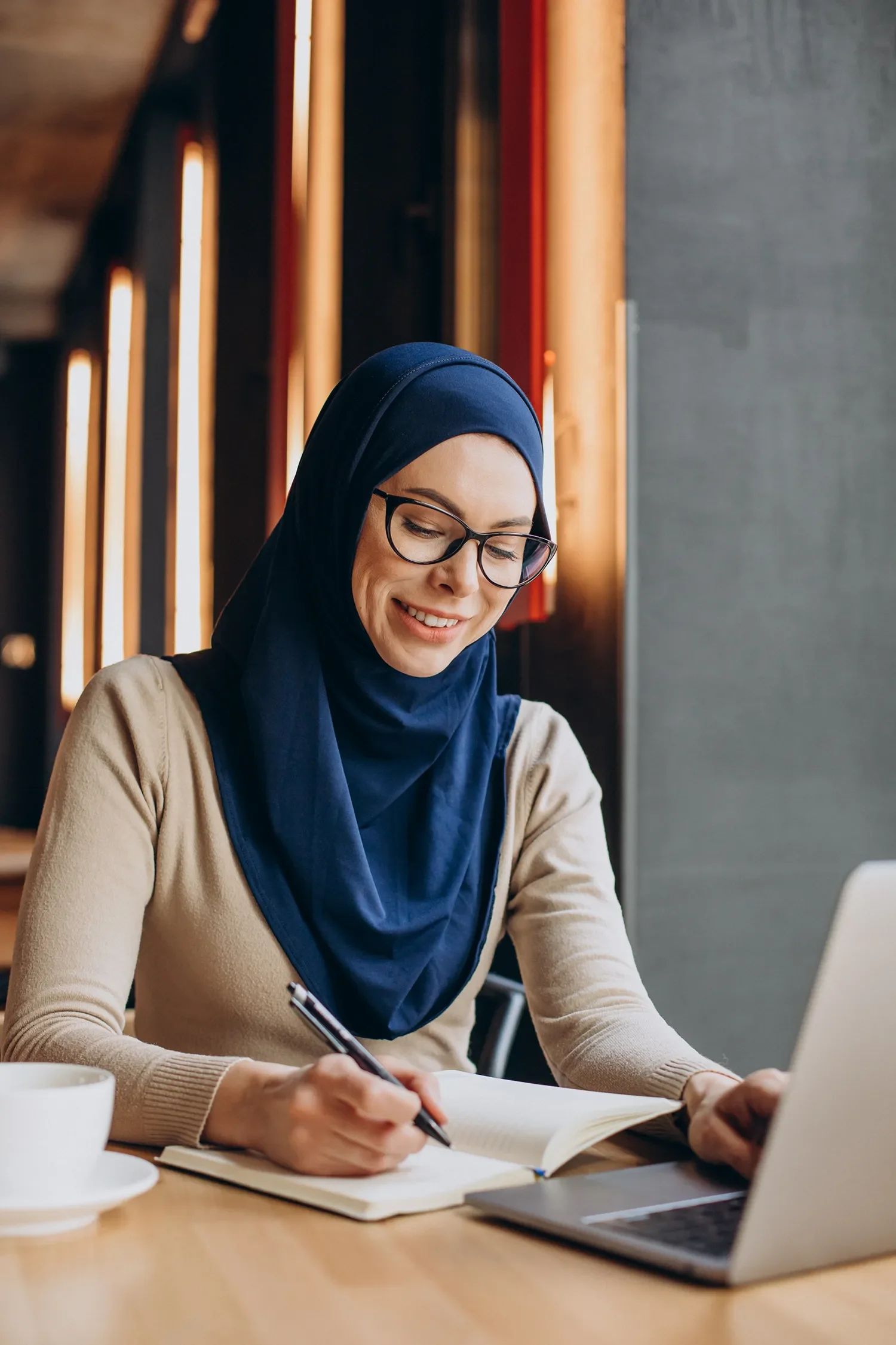 A woman wearing glasses and a navy blue headscarf writes in a notebook at a desk in a cafe or workspace.
