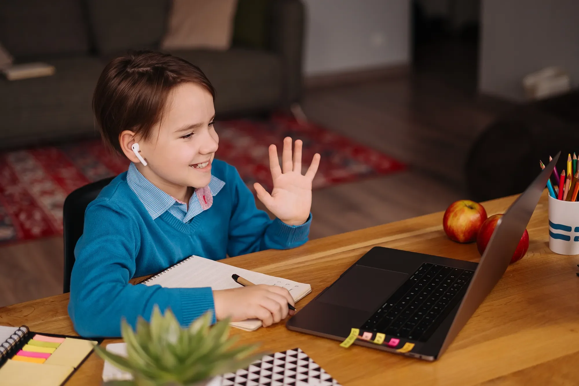 A young boy with brown hair wearing a blue sweater and wireless earbuds, smiling and waving while sitting at a wooden desk with a computer laptop, notebooks, a pen, colorful sticky notes, and apples, in a cozy room with books and furniture in the background.