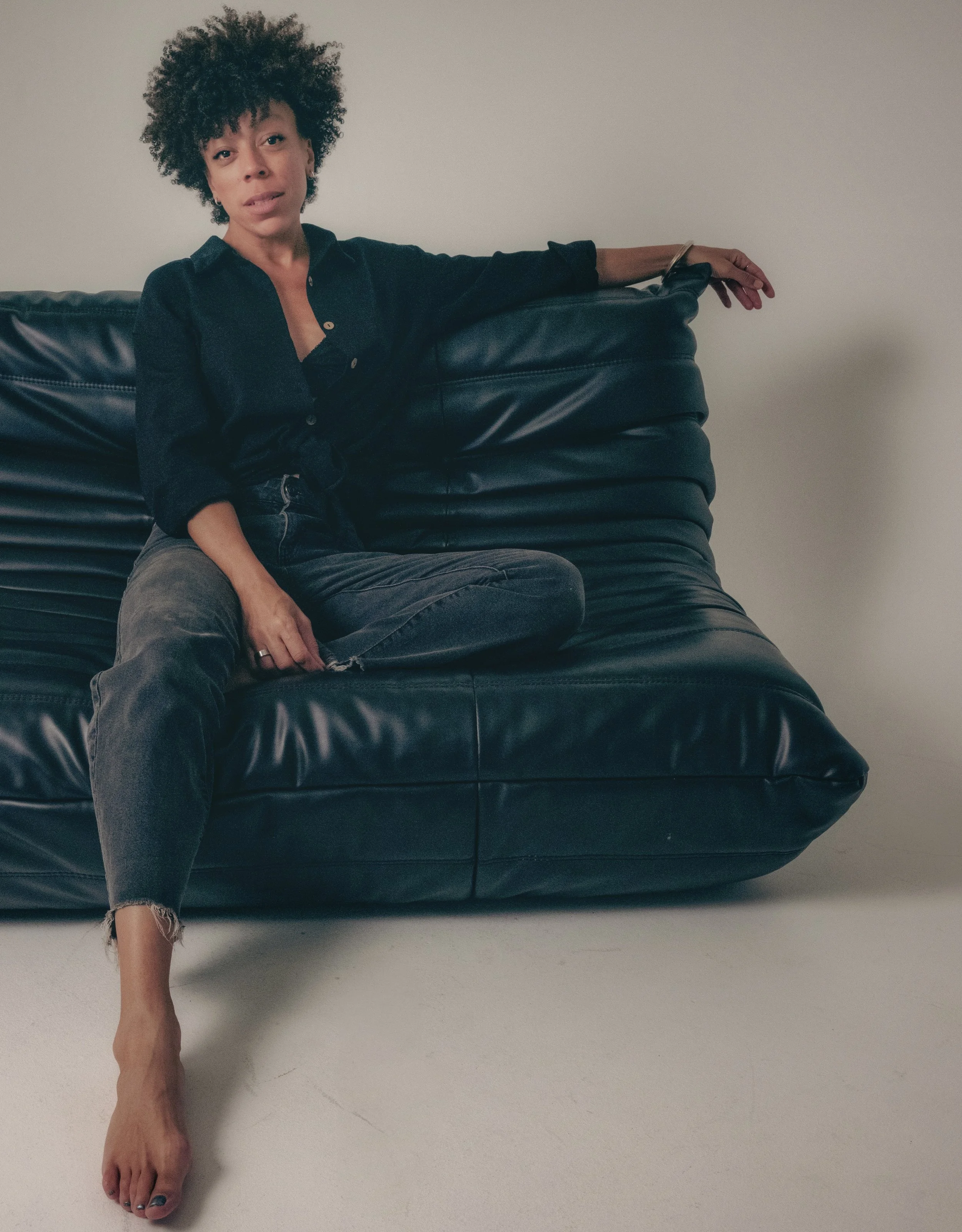 Woman sitting on a black leather couch against a plain wall, dressed in dark clothing and barefoot.