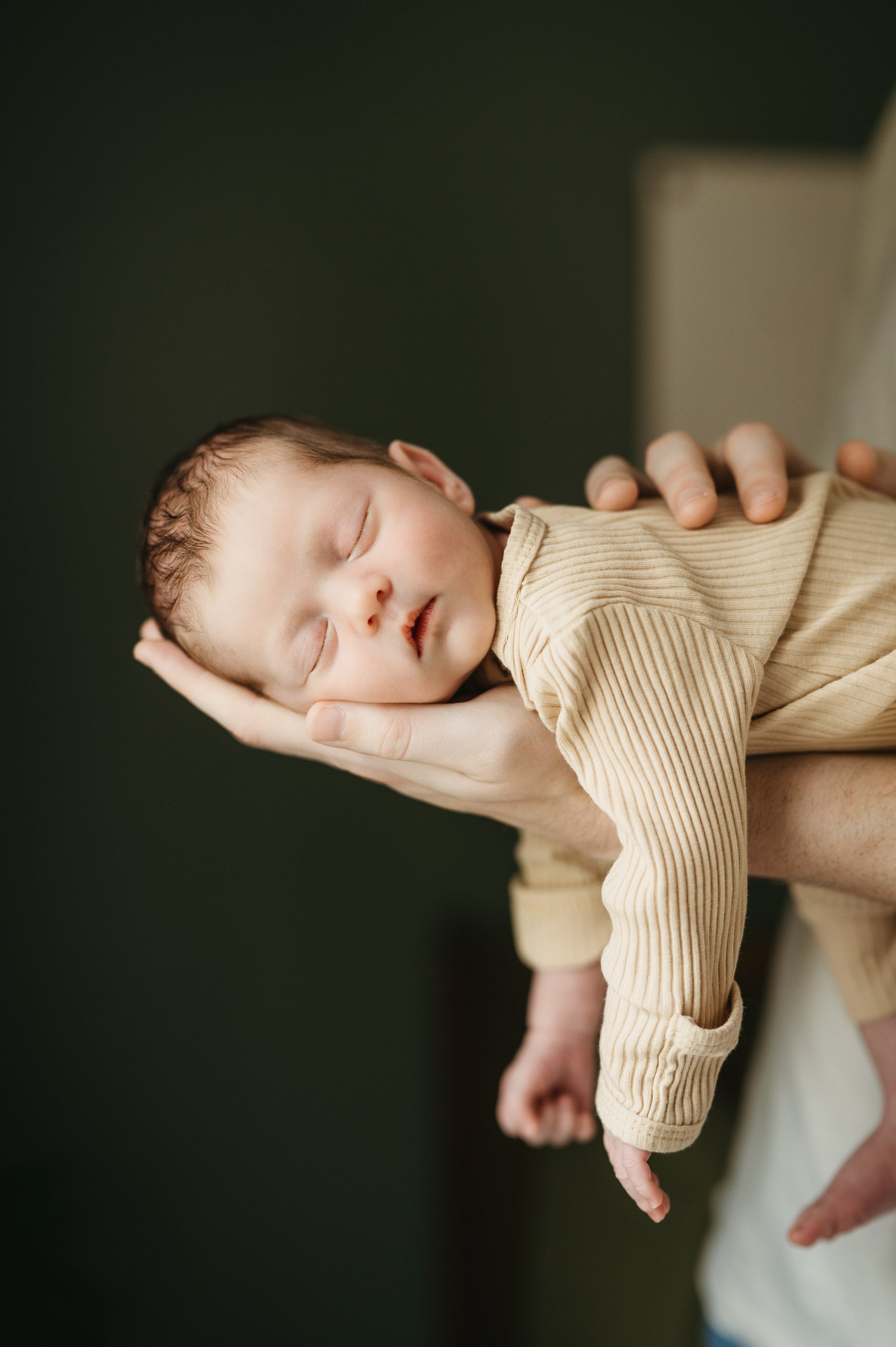Close-up of a sleeping newborn baby being gently held in an adult's hand, resting on its side with eyes closed, a peaceful expression, in a beige ribbed outfit.