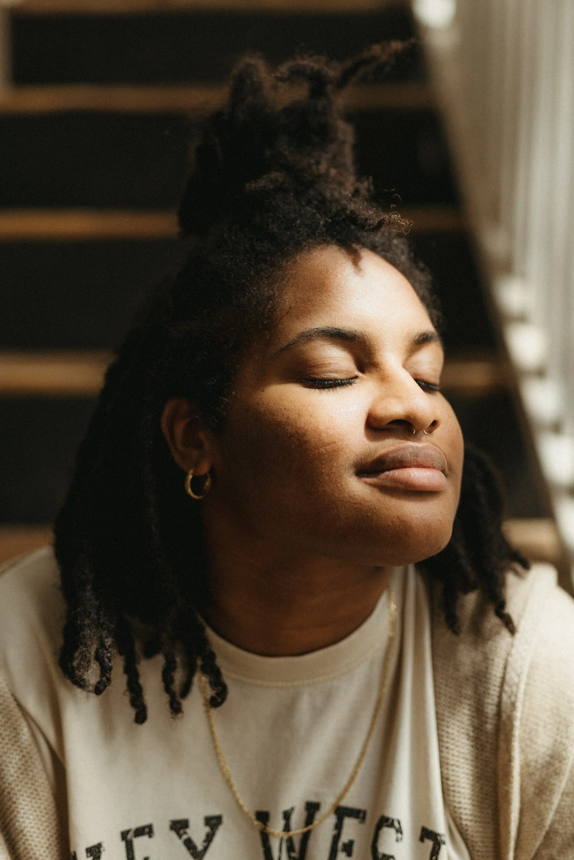 A woman with dark skin and short dreadlocks has closed eyes and a peaceful expression. She is wearing earrings and a necklace, and a light-colored shirt with text partially visible. She is sitting indoors near a window.