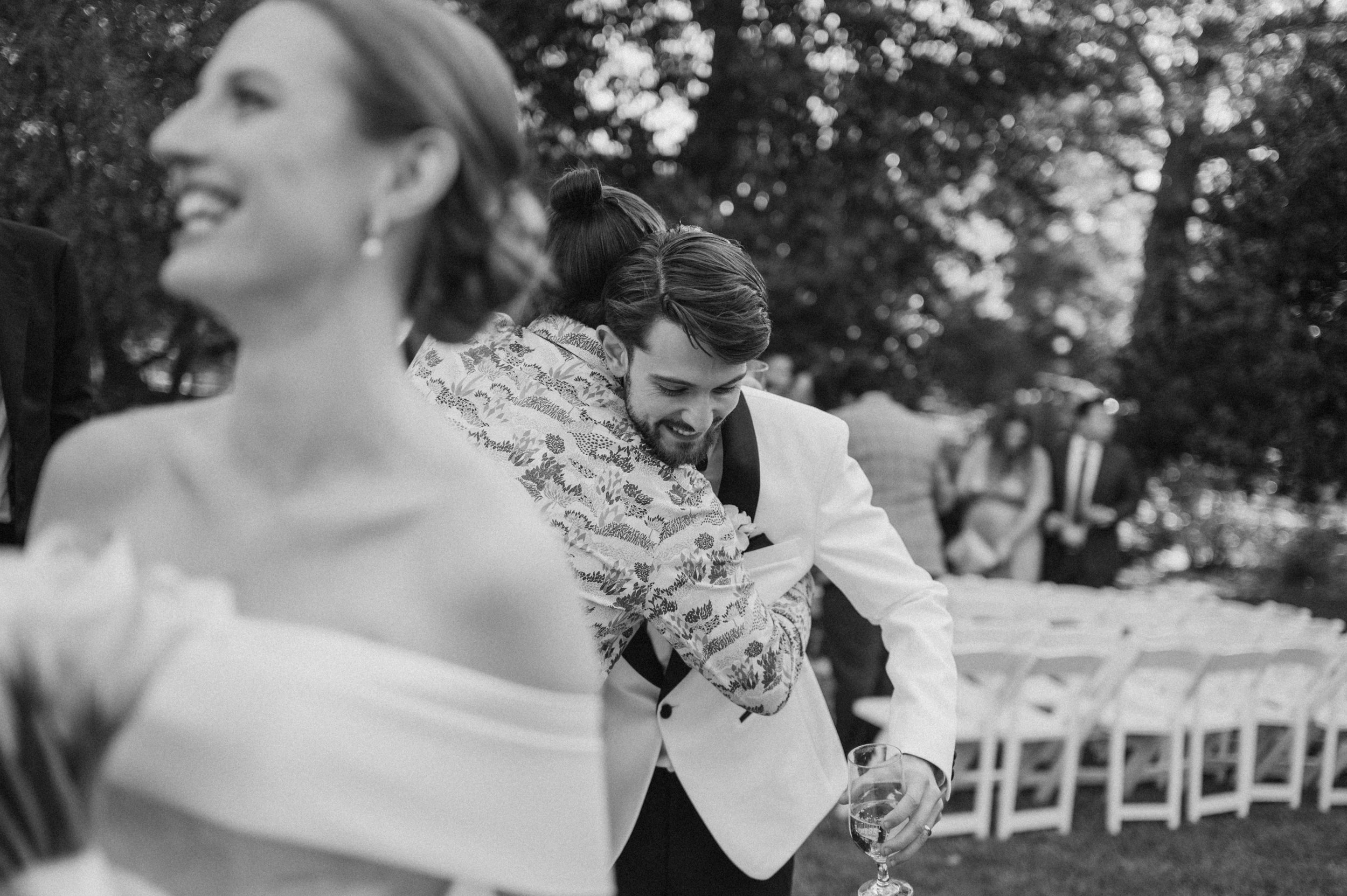 A black and white photo of a wedding reception outdoors, with a bride in a strapless dress in the foreground and two guests hugging in the middle ground, one in a white jacket holding a glass of wine. Several chairs are visible in the background with trees and people.
