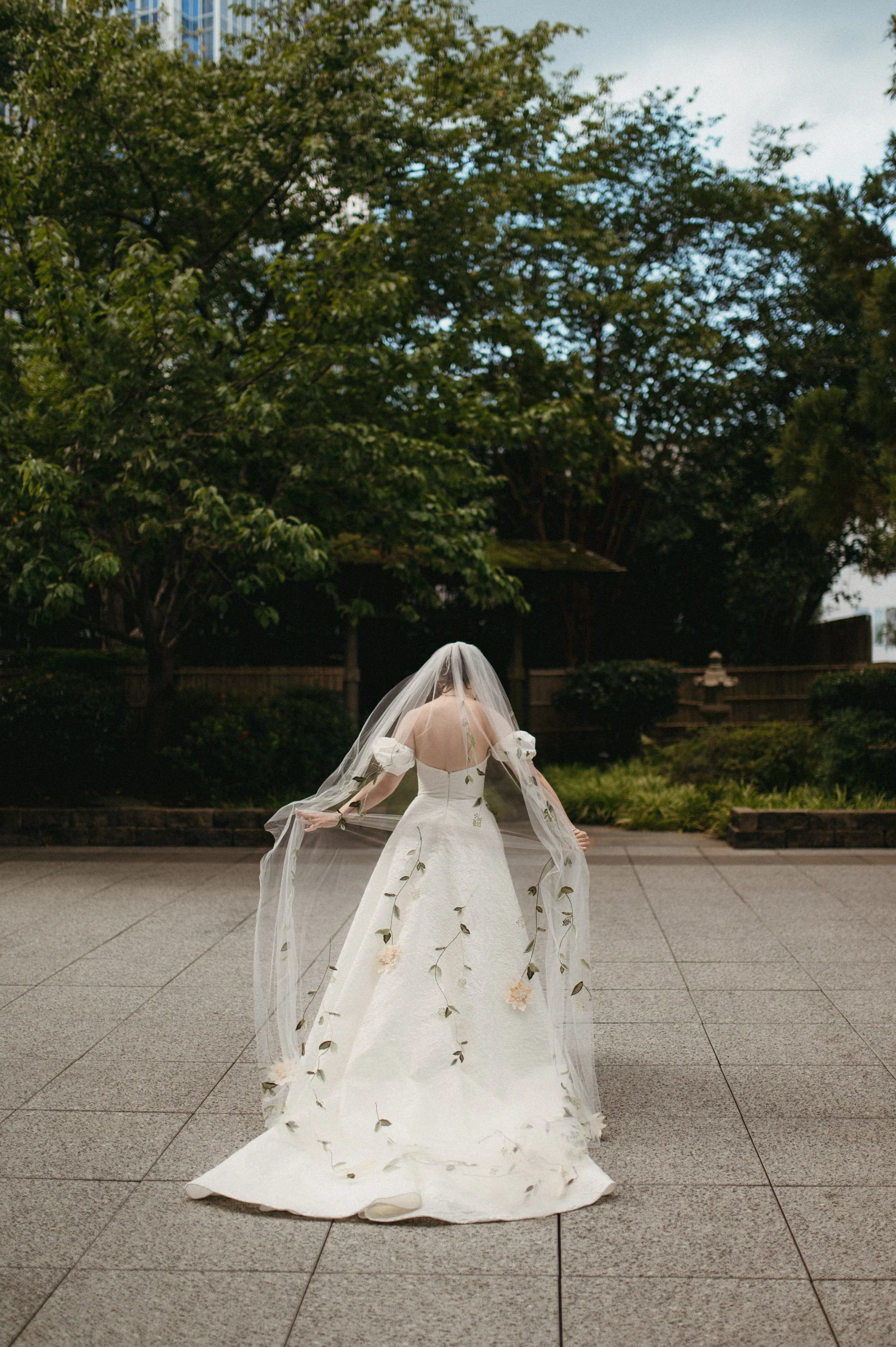 A bride in a white wedding dress with floral embroidery, holding her veil, standing on a paved outdoor area with trees and a fence in the background.