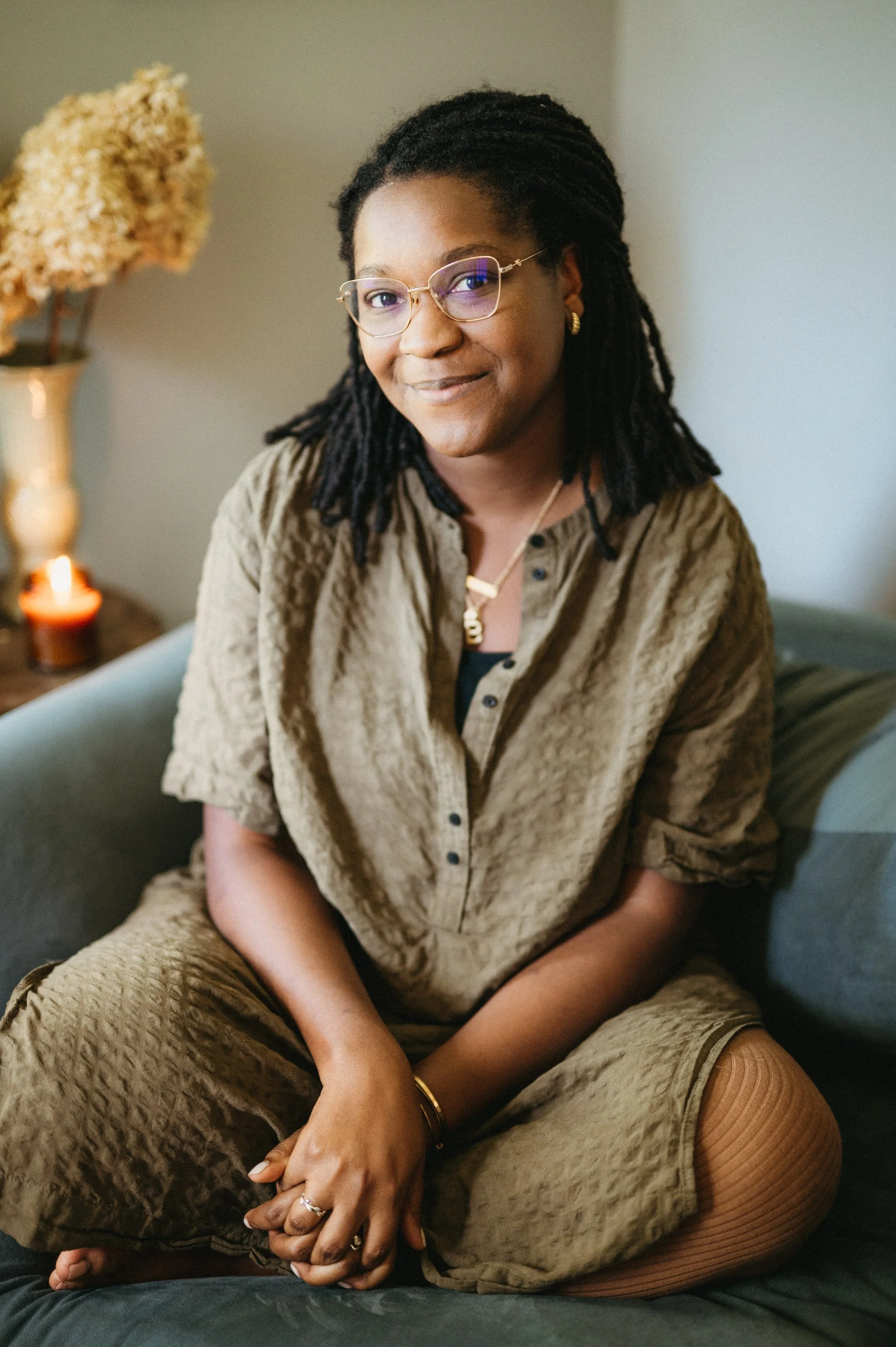 A woman with glasses and dreadlocks sitting on a couch, smiling at the camera. She is wearing a textured, loose-fitting, brown shirt and matching pants, with jewelry including rings, a necklace, and earrings. In the background, there is a vase with d