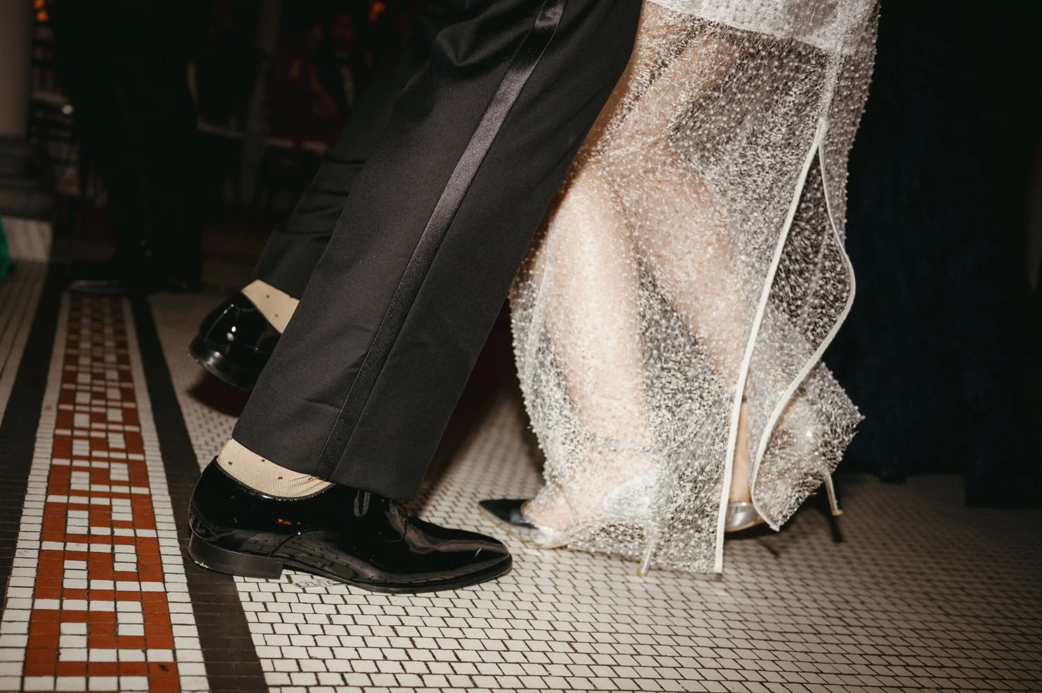 Close-up of a couple's legs and feet, with the man wearing black dress shoes and the woman wearing silver high heels, seated on a mosaic-tile floor.