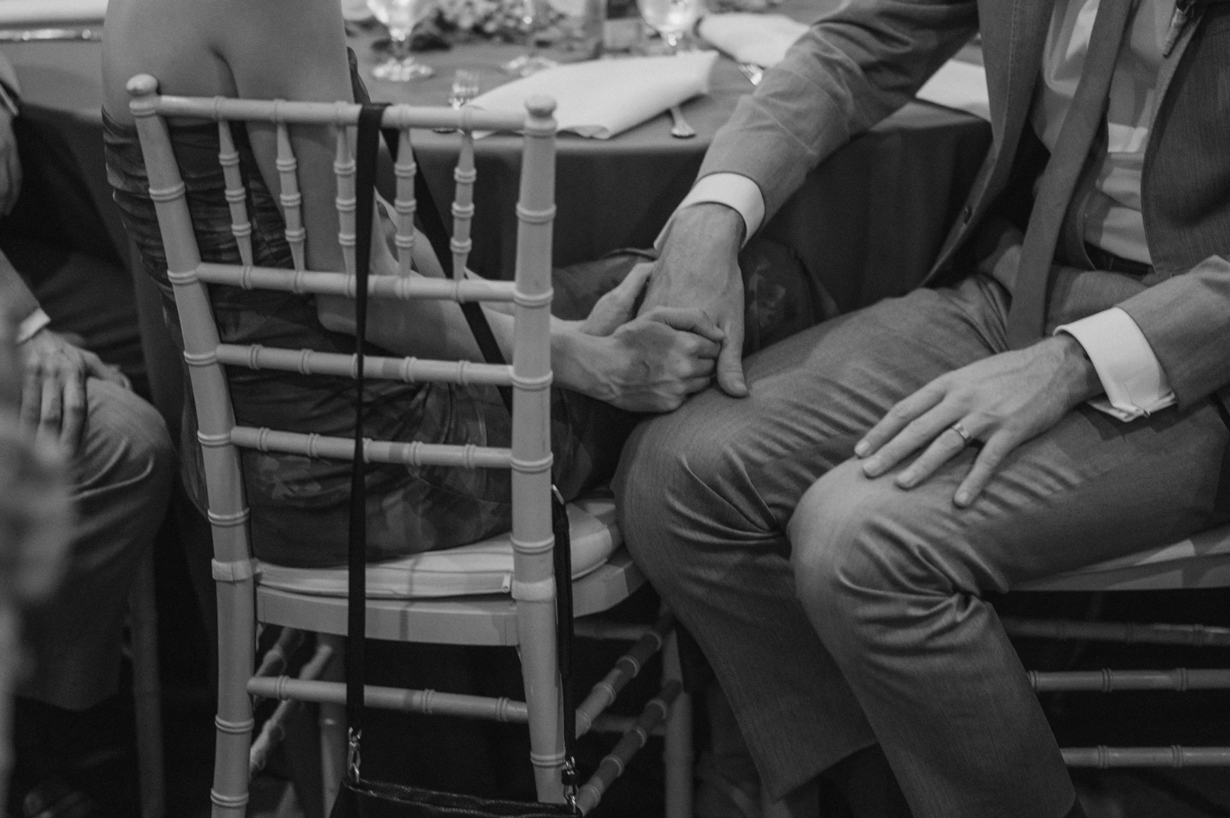 A black and white photo showing two people holding hands at a formal event, seated at a table with utensils and glasses.