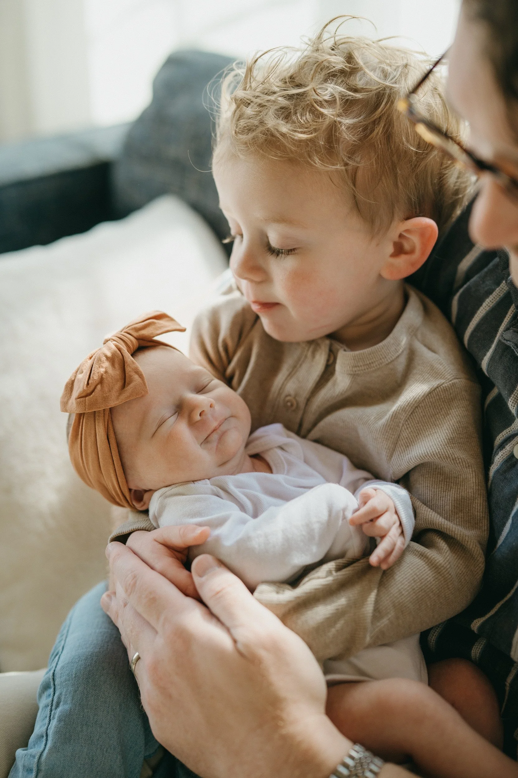 A young boy gently holding and looking at a sleeping newborn girl, who is wearing a headband with a bow.