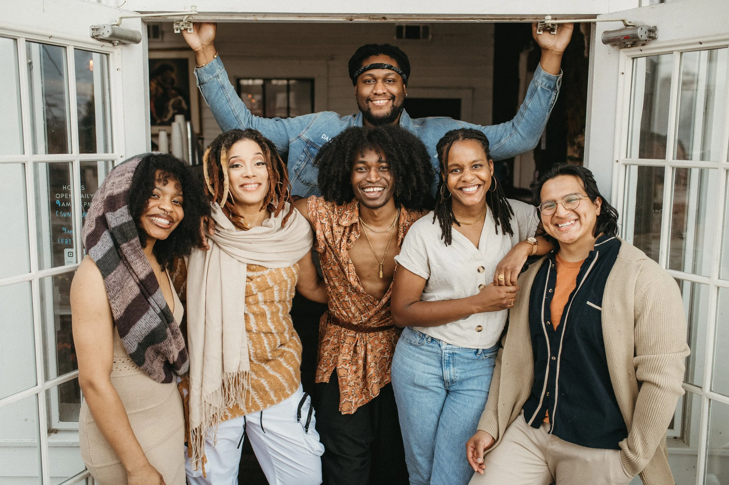 Group of six diverse happy friends posing together at a party or gathering, standing in front of an open door, smiling at the camera.
