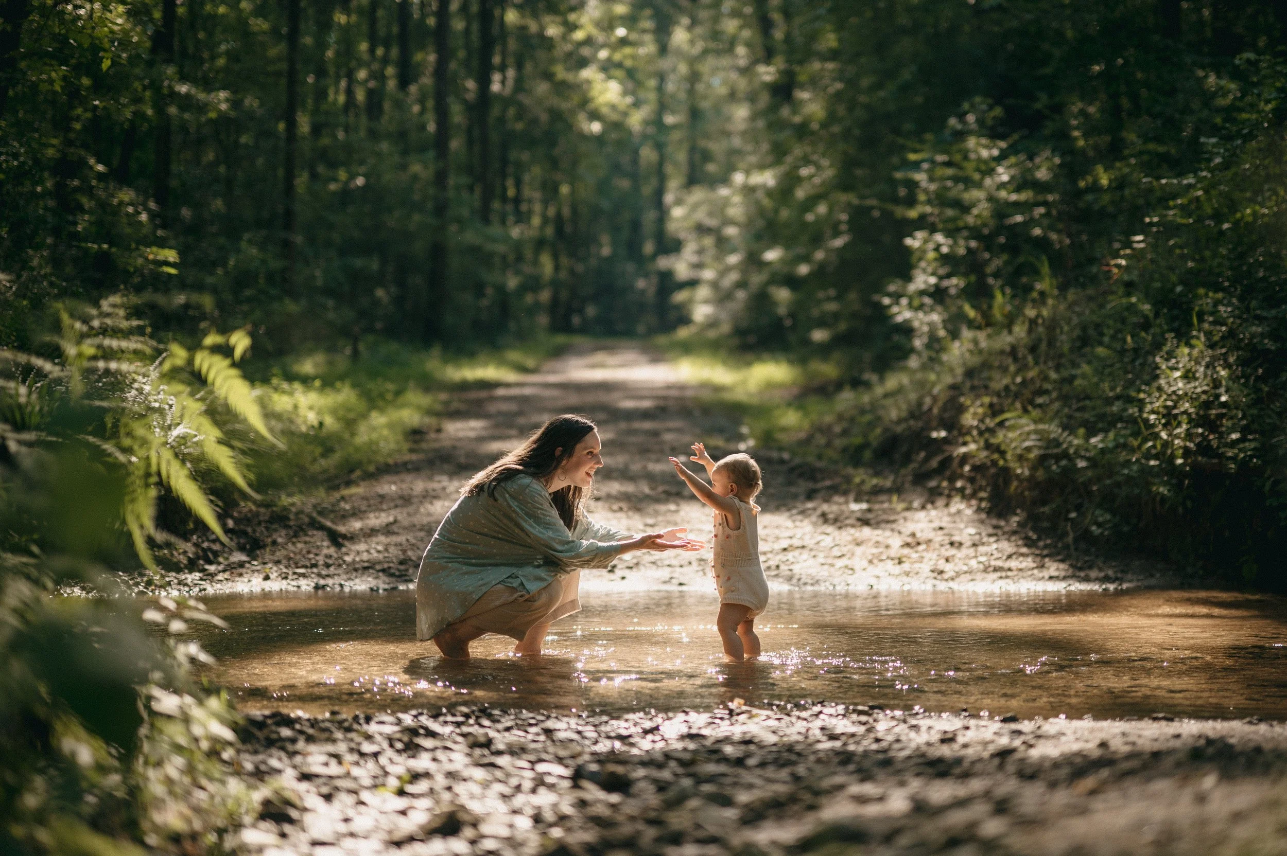 A woman and a young child playing in a shallow creek in a forest with sunlight filtering through the trees.