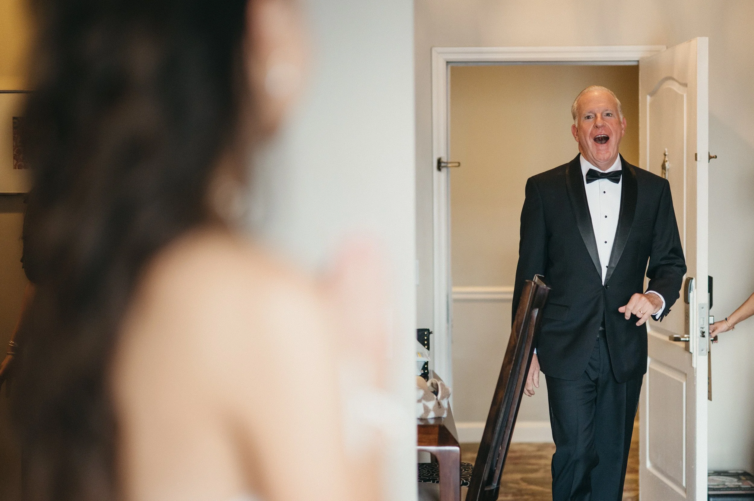 An elderly man in a tuxedo with a bow tie, smiling and walking into a room, possibly during a wedding or formal event.