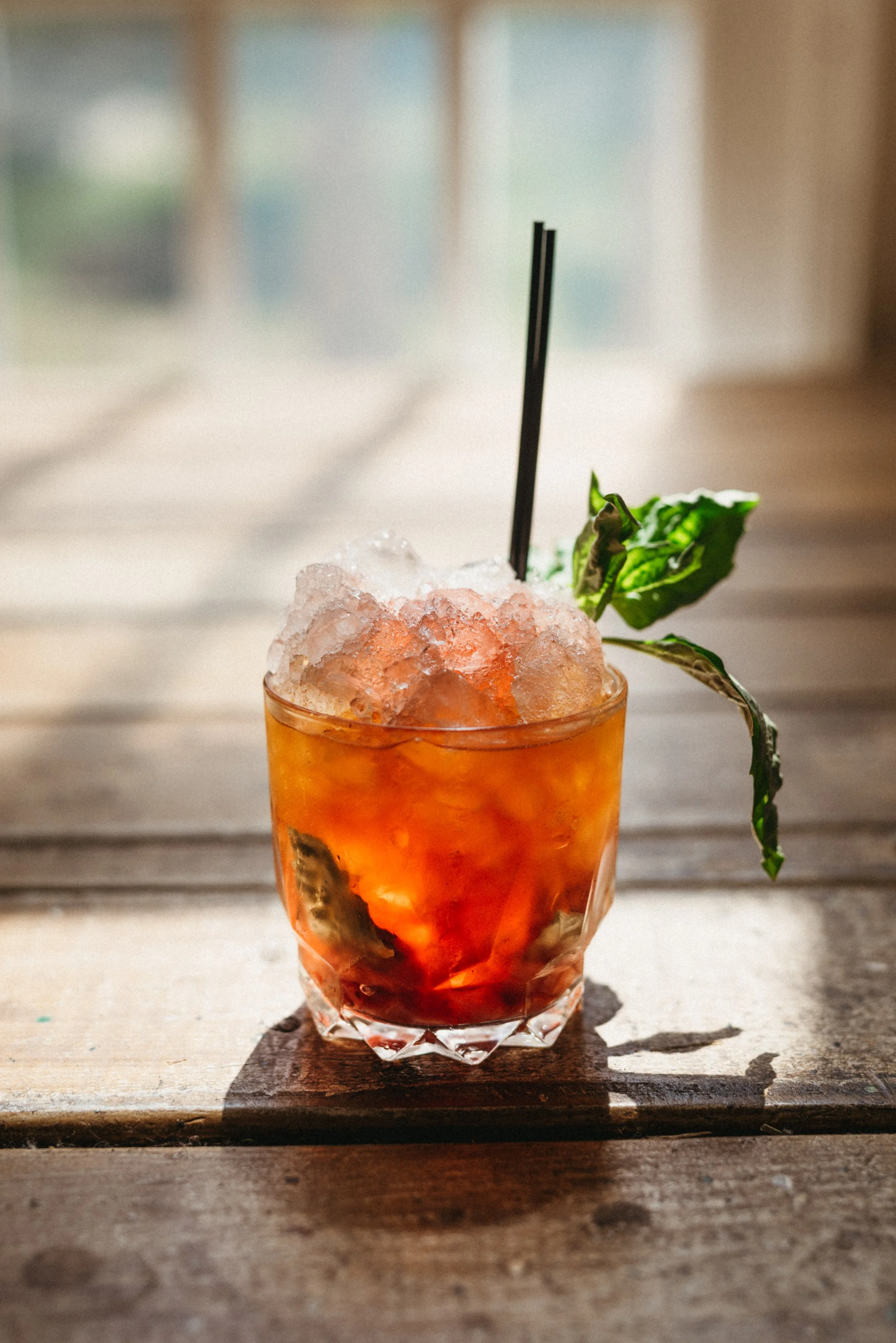 A glass of iced cocktail garnished with a basil leaf and a black straw, placed on a wooden table with sunlight.