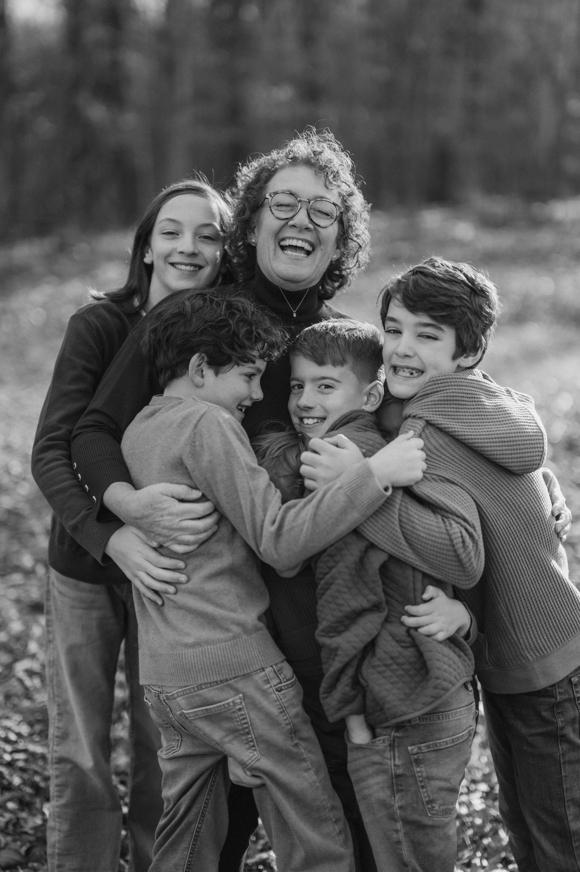 A black and white photo of a woman and five children hugging and smiling outside in a wooded area.