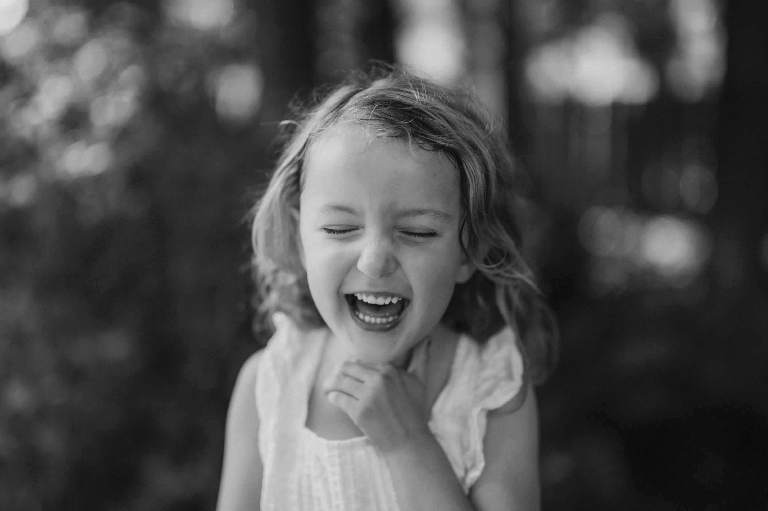 Black and white photo of a young girl laughing with her eyes closed outside, surrounded by blurred trees.