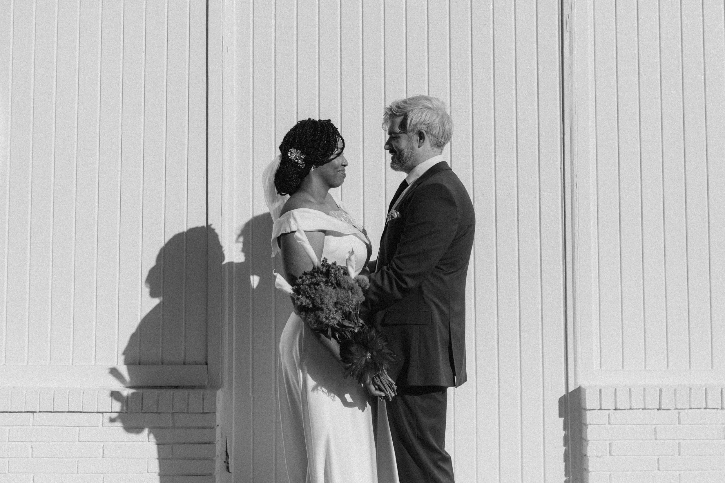 A black-and-white photo of a couple in wedding attire standing close together, gazing into each other's eyes, against a background of a light-colored wooden wall. The bride holds a bouquet and has fashionably styled hair adorned with a decorative hai