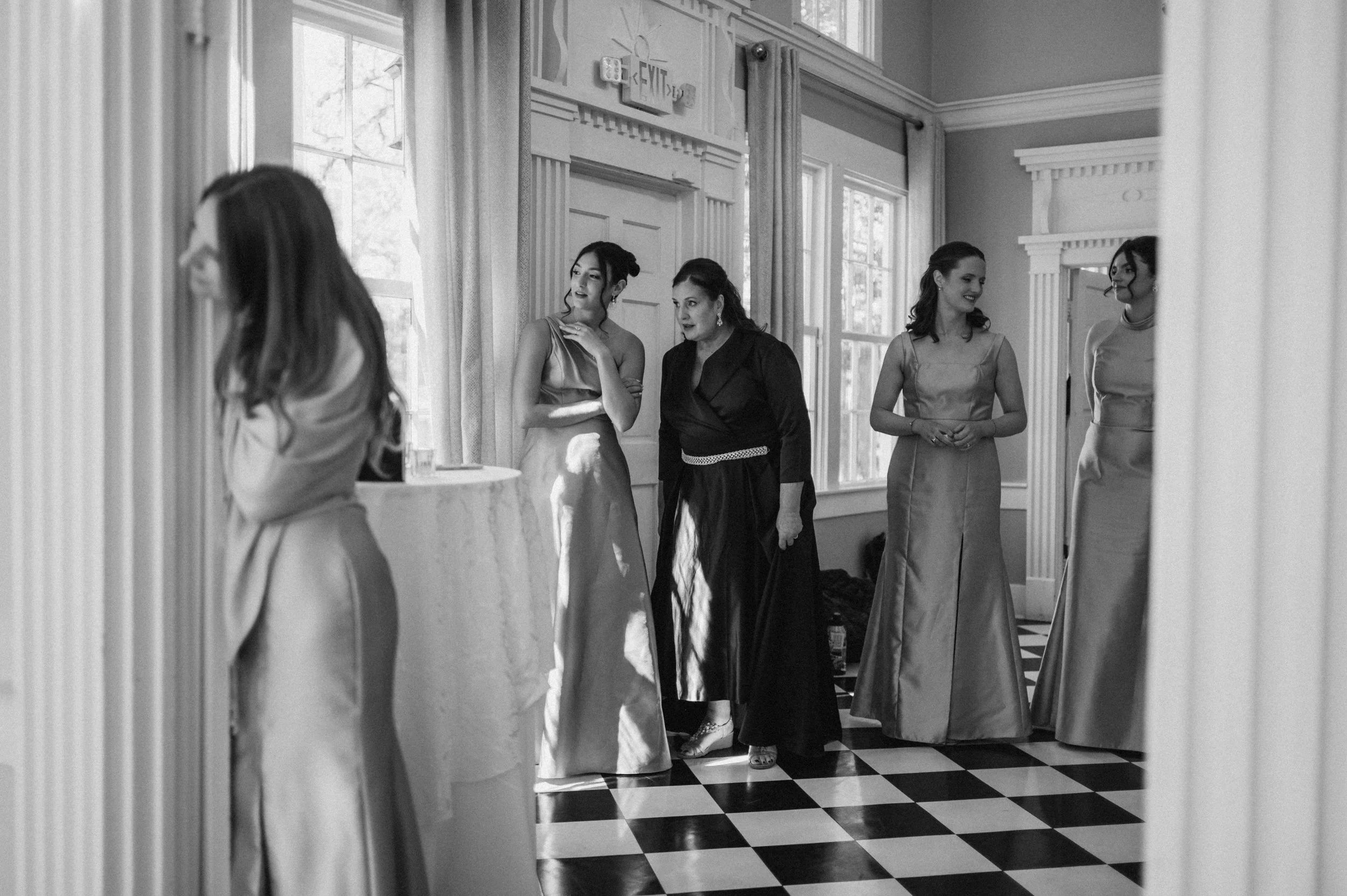 Women in elegant dresses standing and conversing in a sunlit room with checkered floor, large windows, and decorative moldings.