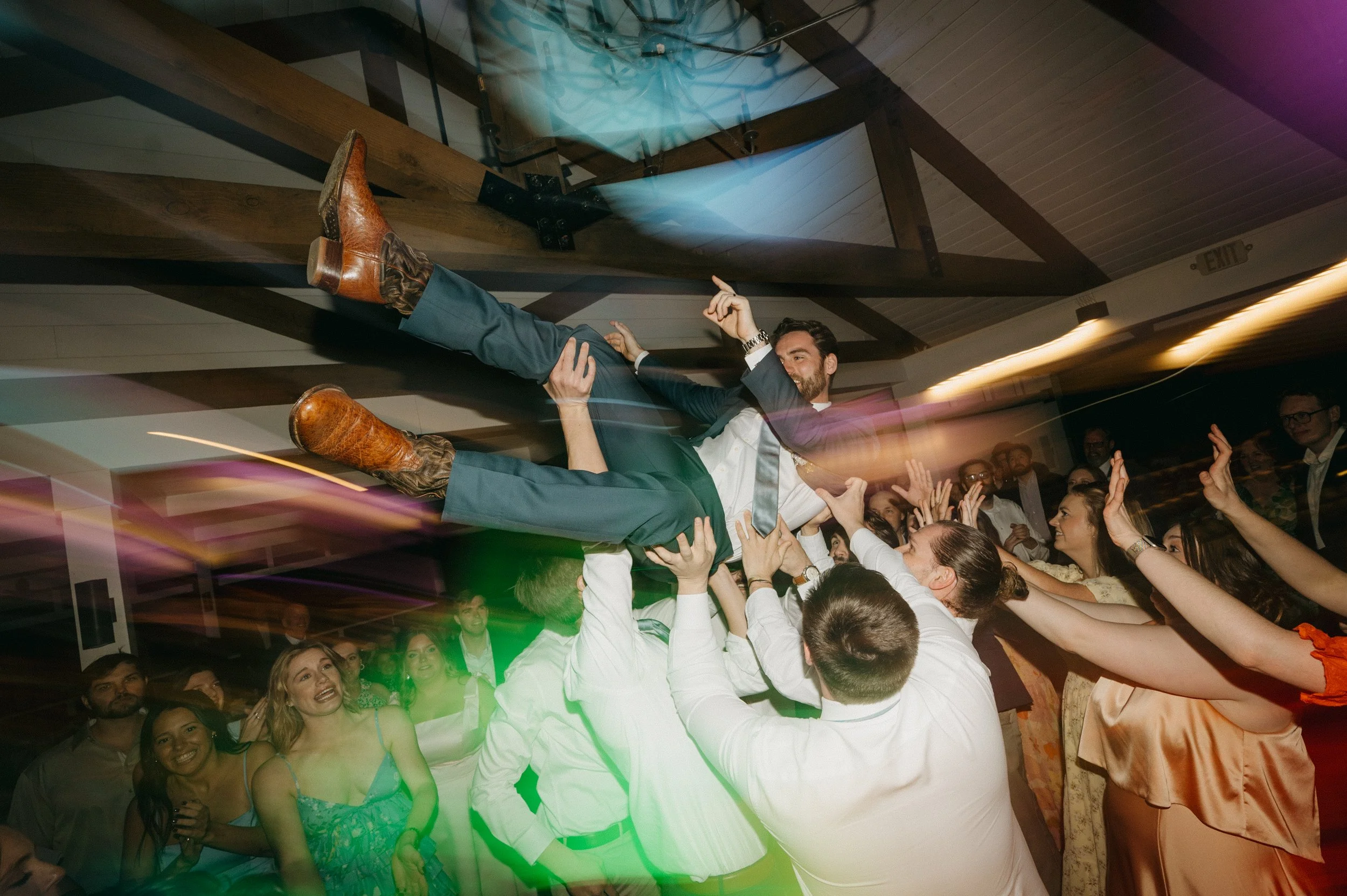 A man in a suit being lifted and crowd surfing at a lively event or party, with people reaching up and smiling, colorful lights and motion blur.
