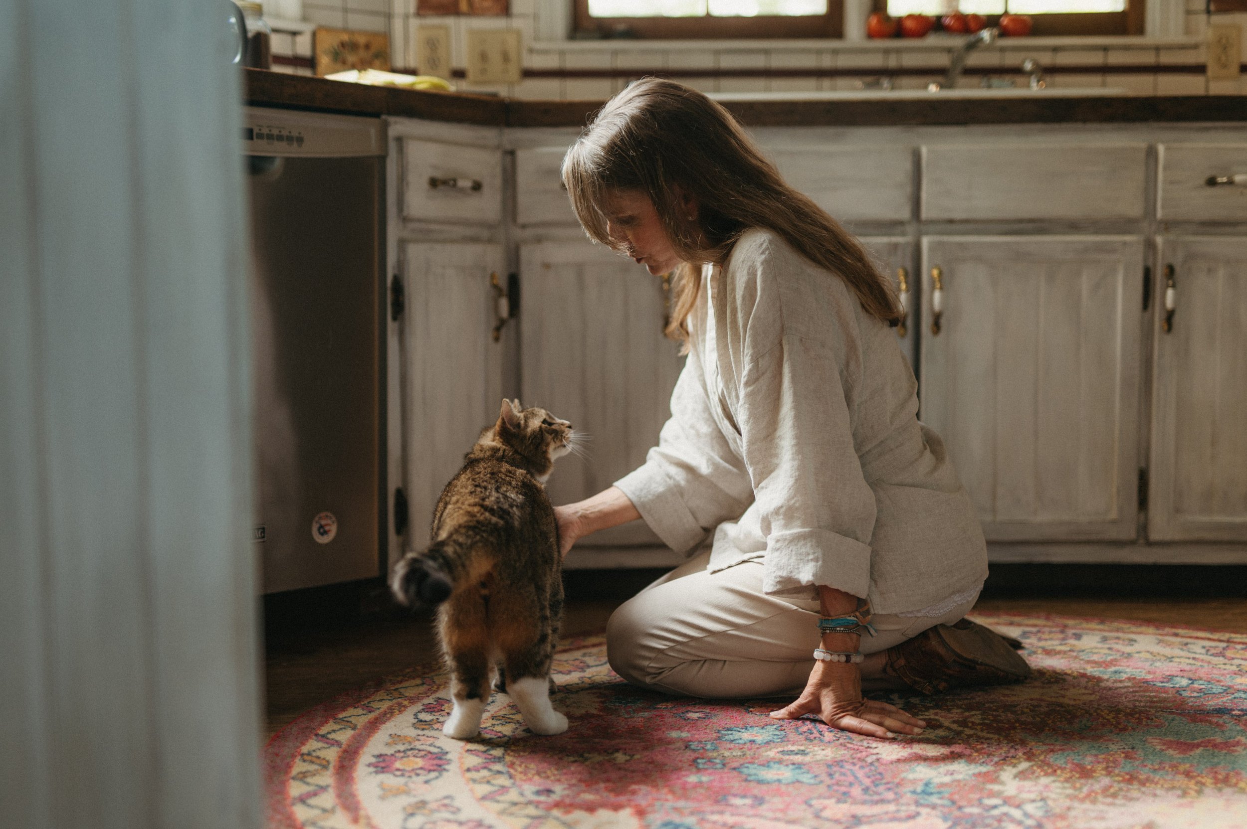 A woman is kneeling on a colorful rug in a kitchen, petting a small tabby kitten.