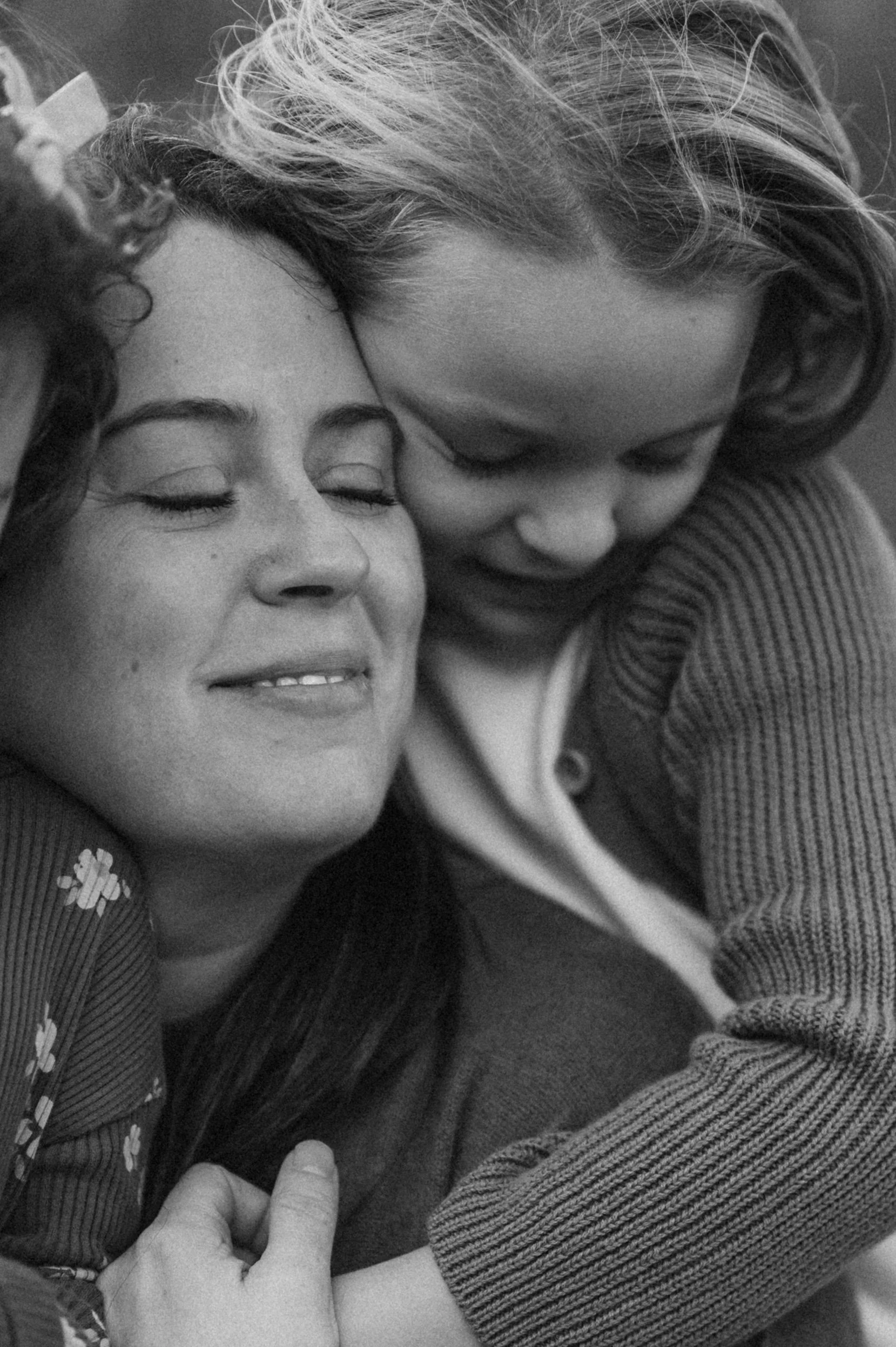 A black and white close-up photograph of a woman and a young girl sharing an embrace, with both smiling and eyes closed.