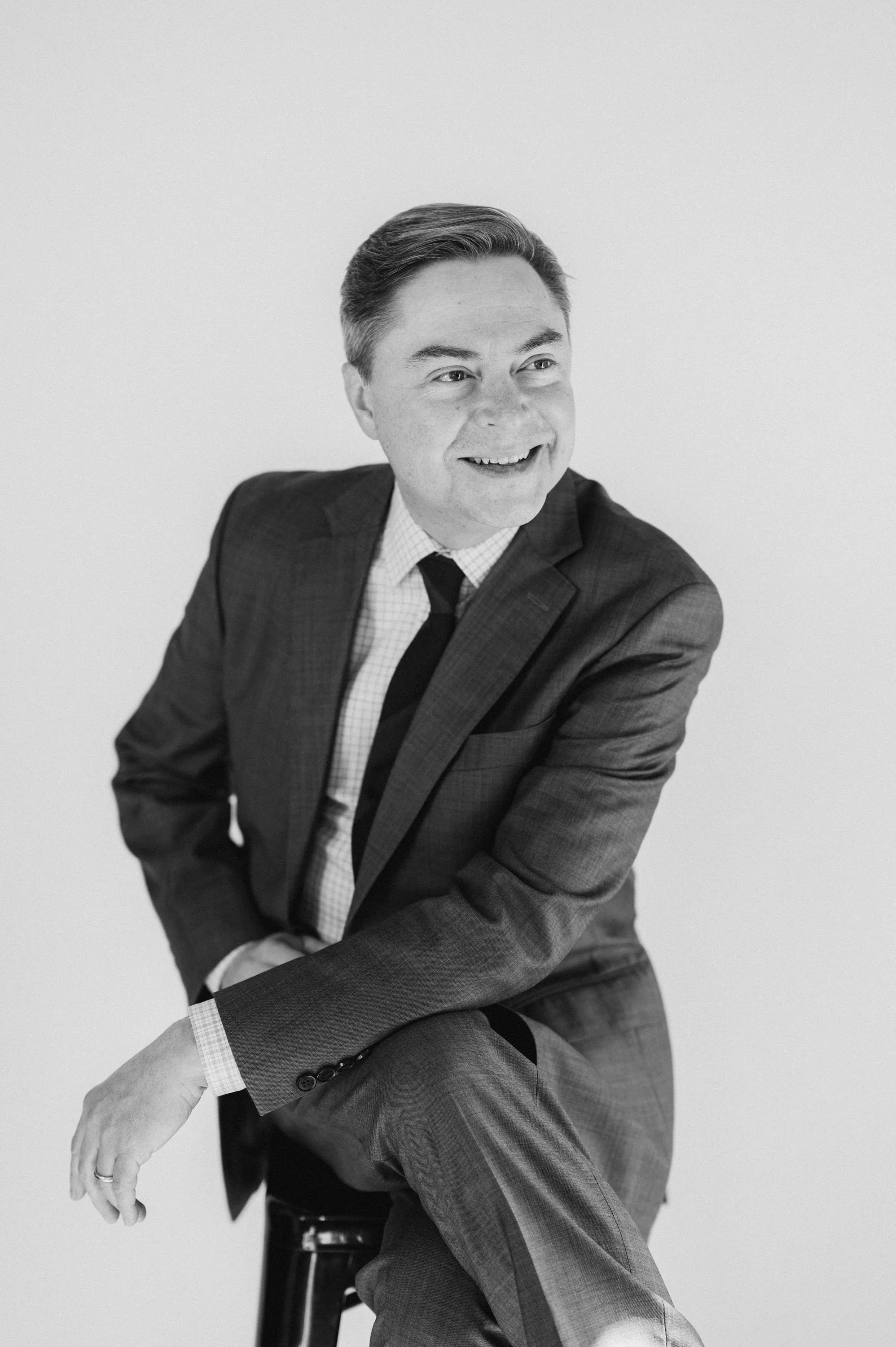 Black and white photo of a man in a suit sitting on a stool, smiling and looking to the side against a plain background.