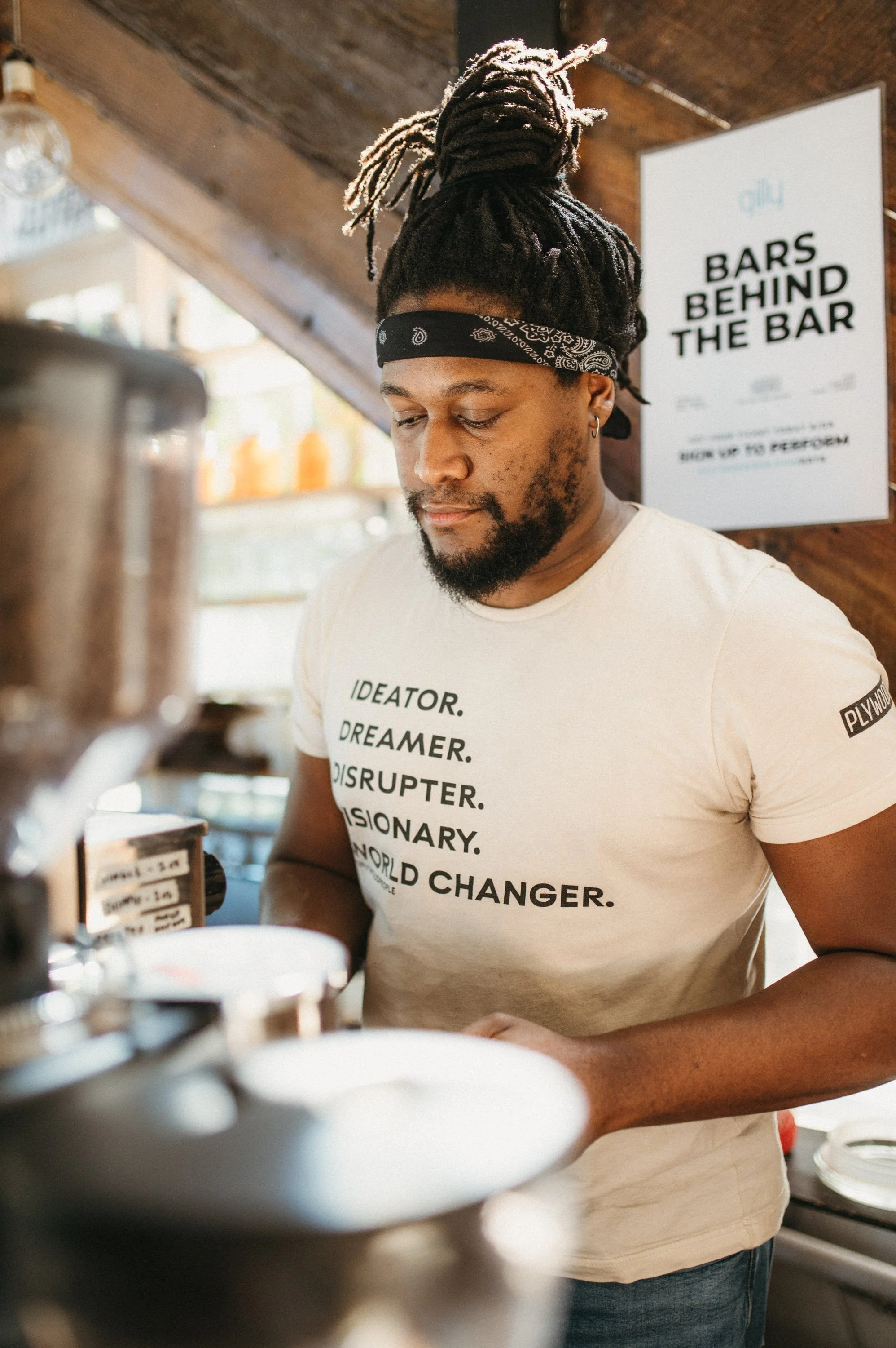 A man with dreadlocks tied up, wearing a black bandana and a cream T-shirt with words like 'IDEOATOR', 'DREAMER', 'DISRUPTER', 'VISIONARY', and 'WORLD CHANGER' printed on it, working behind the bar in a cafe or bar.
