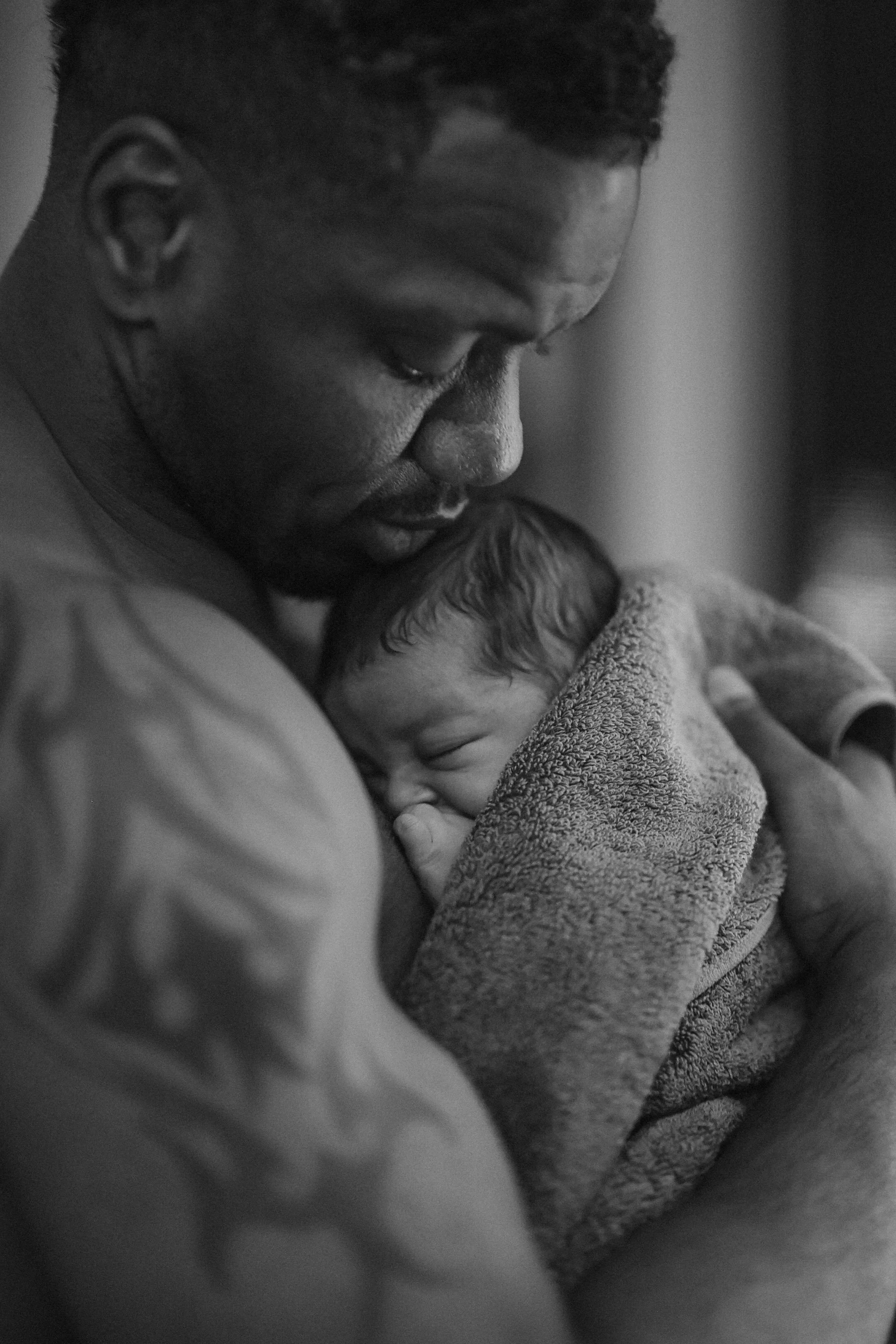 A man holding a sleeping infant close to his chest, with the man's head bowed down touching the baby's head, in a tender moment.