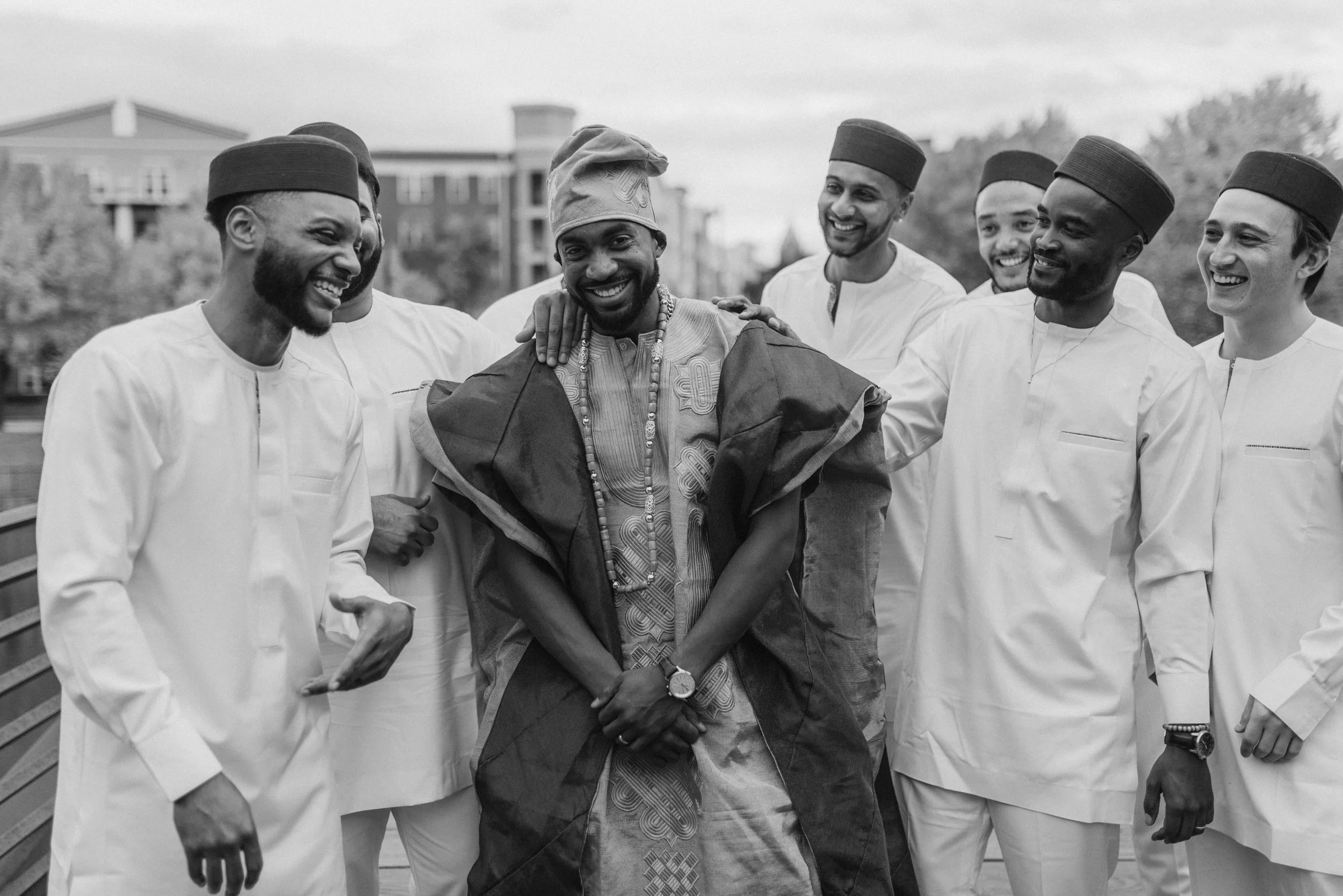 A group of seven men, some wearing traditional Nigerian attire and others in white chef uniforms, are smiling and laughing together outdoors.
