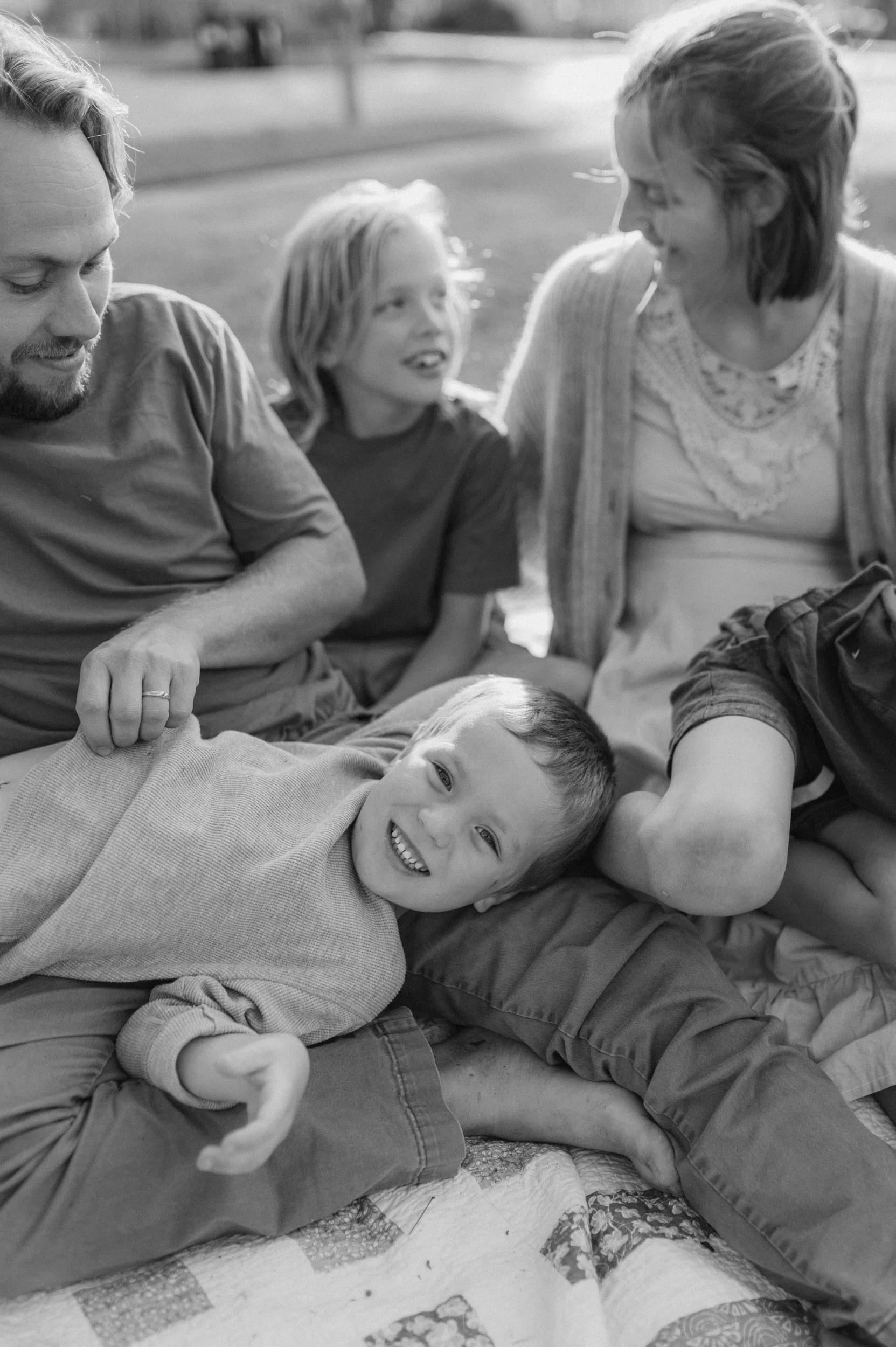 A family of four sitting on a blanket outdoors, smiling and enjoying each other's company.