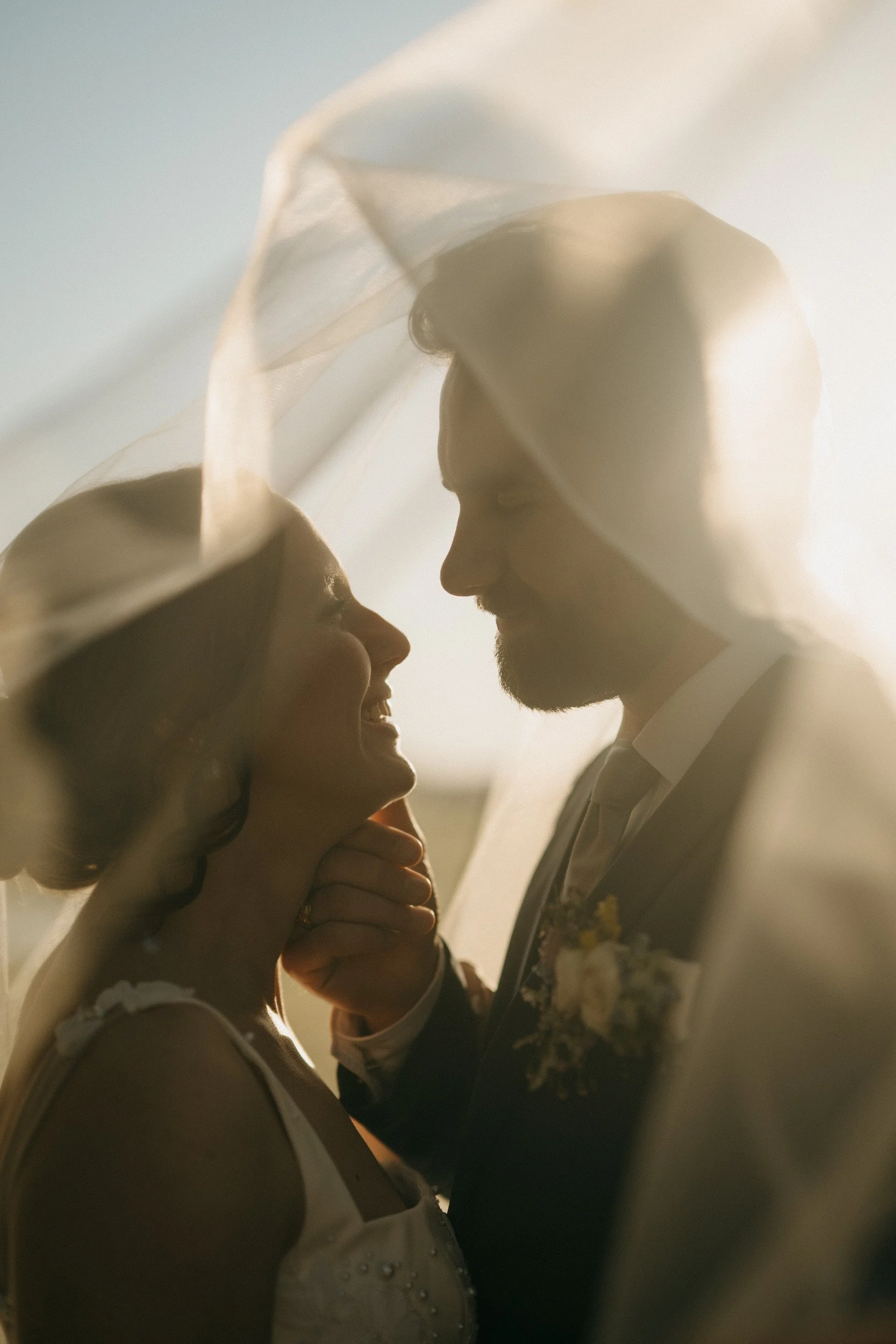 A bride and groom on their wedding day, sharing an intimate moment under a soft veil with warm sunlight.
