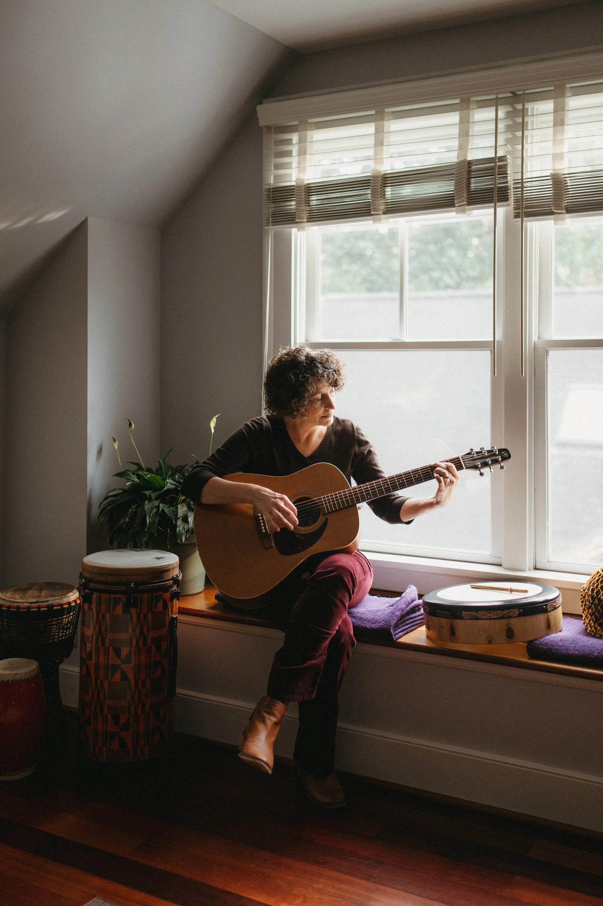 Woman sitting on a window seat playing an acoustic guitar near a large window with blinds, surrounded by musical drums and a potted plant.