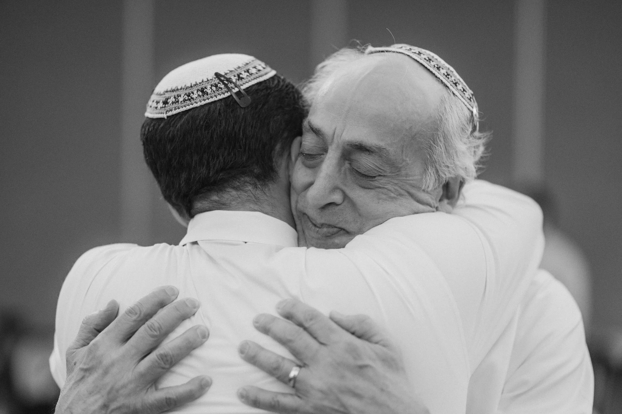 Two men wearing yarmulkes embrace tightly in a heartfelt hug.