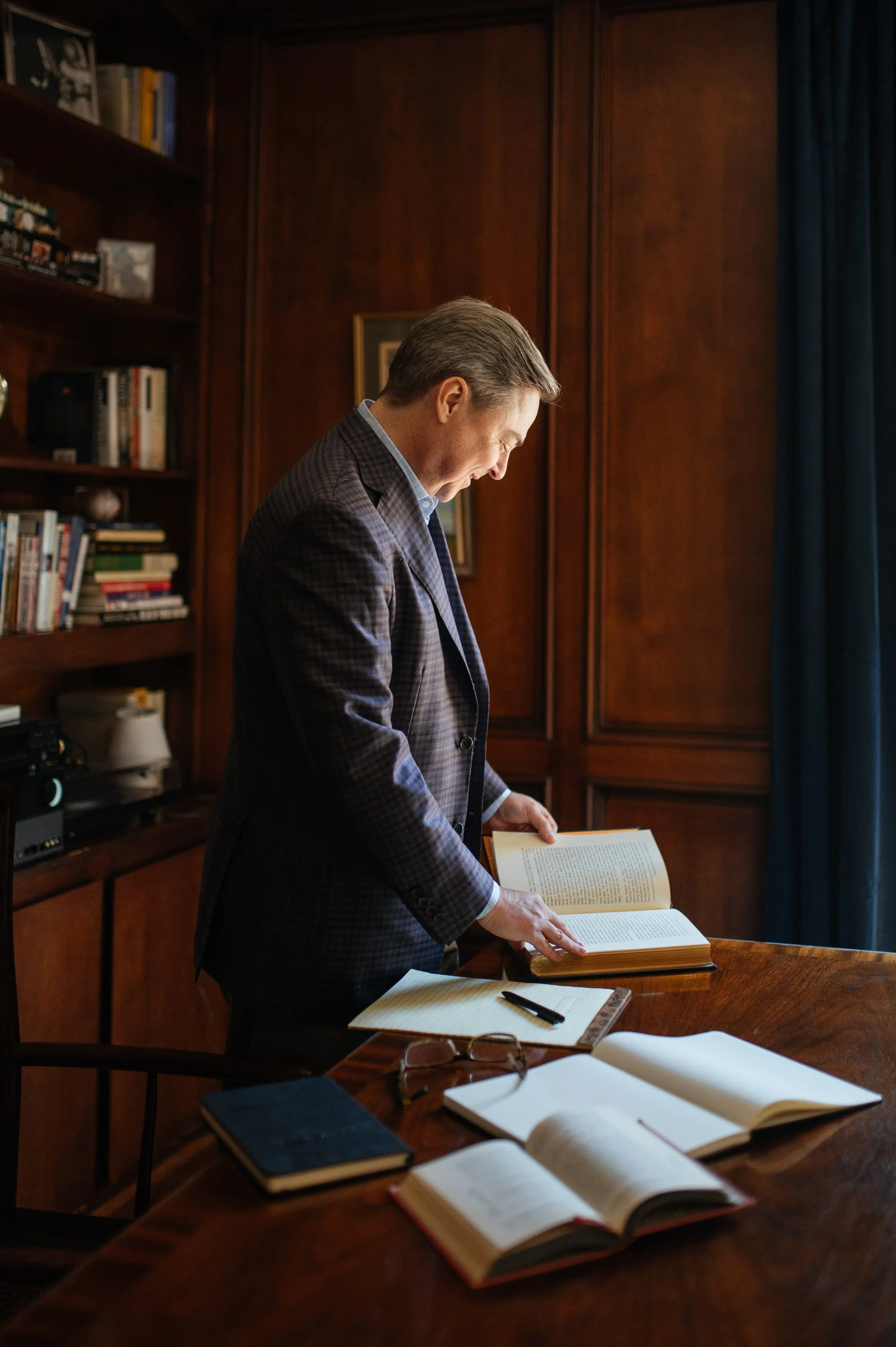 A man in a gray blazer standing at a wooden table, reading a large open book in a room with wooden walls, bookshelves, and a window with blue curtains.