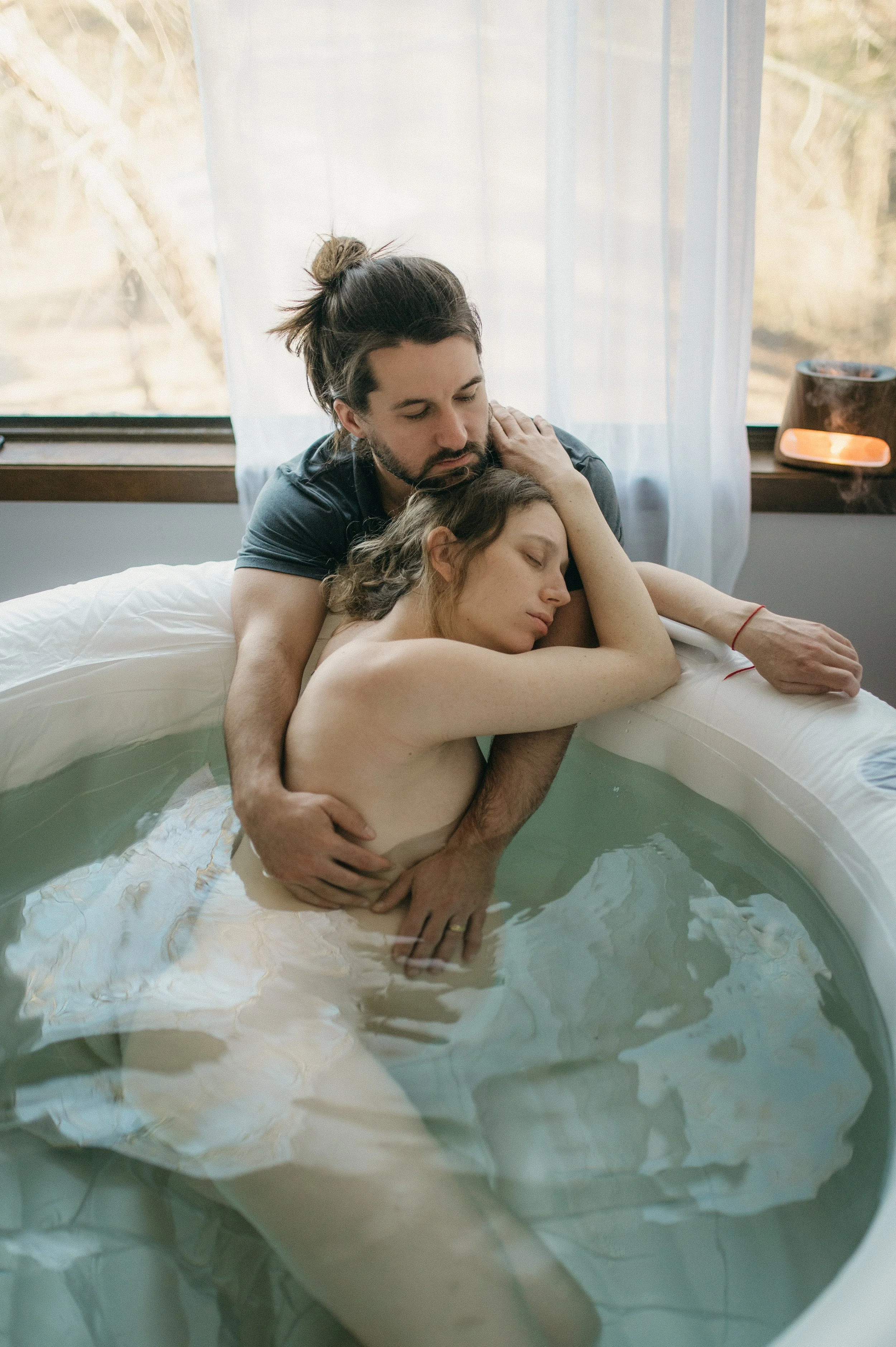 A man and a woman sharing an intimate moment in a hot tub, with the man embracing the woman as she rests her head on his shoulder. They appear to be in a cozy, softly lit room with sheer curtains and a window in the background.
