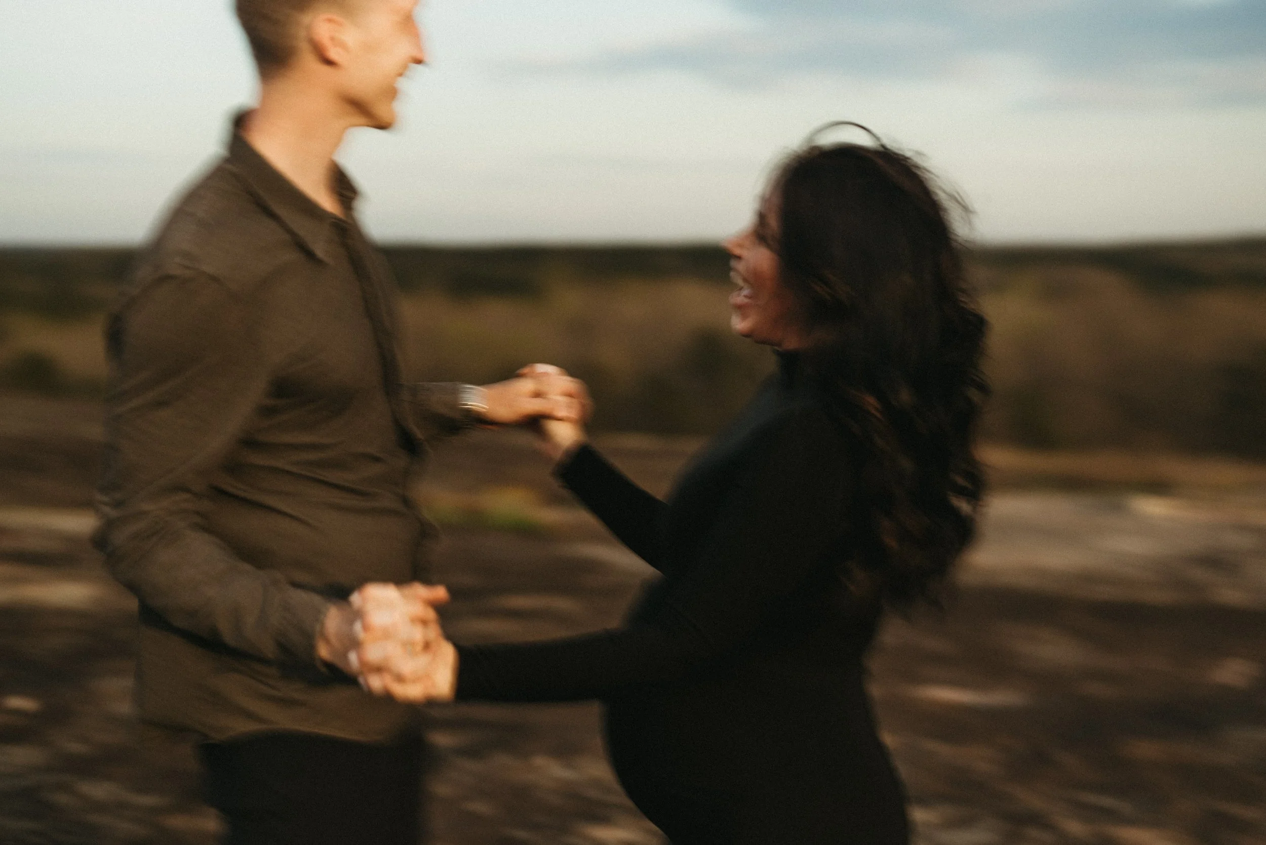 A man and a woman holding hands and dancing outdoors in a blurred landscape at sunset.