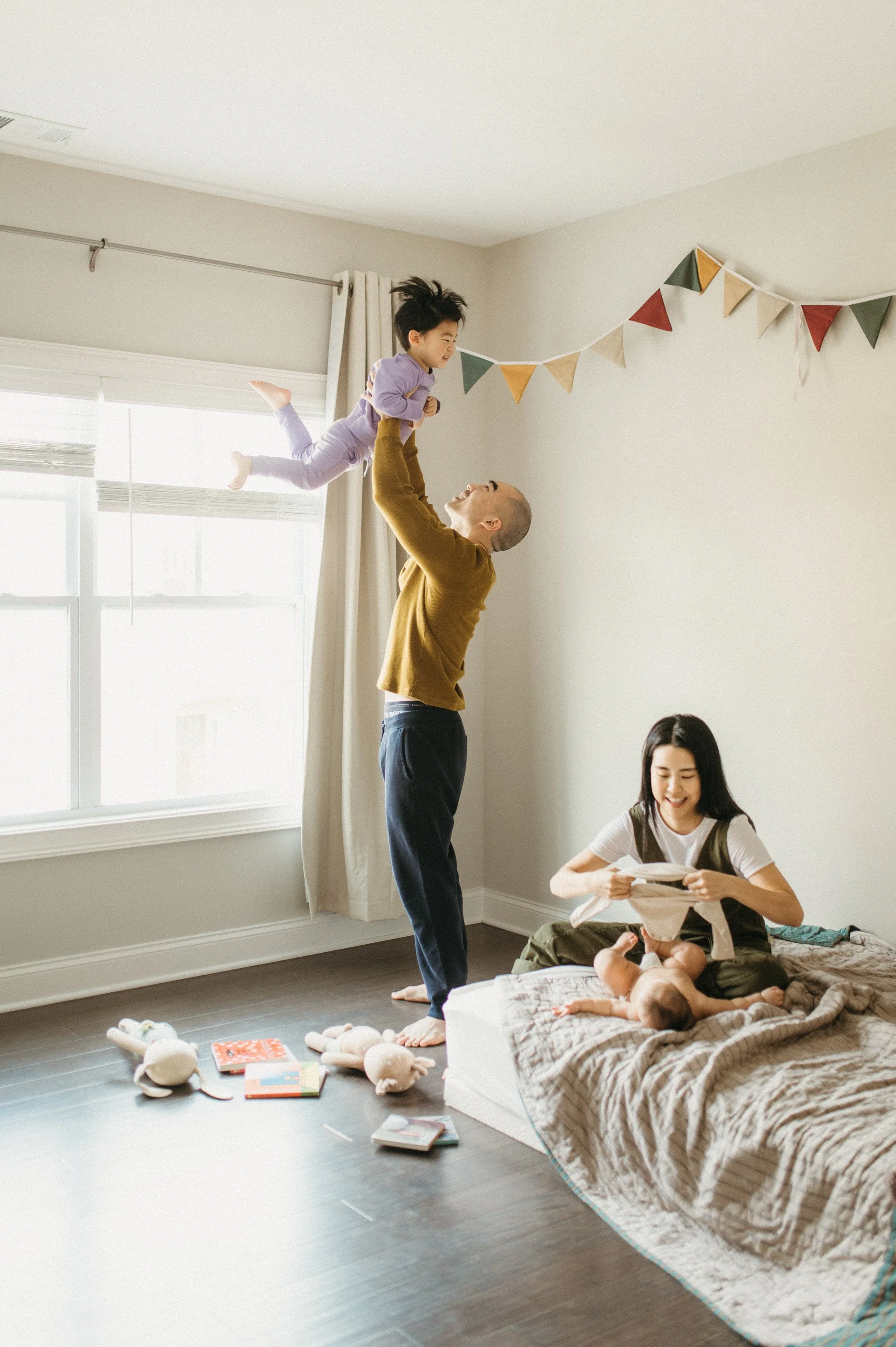 A family enjoying a playful moment in their bedroom, with a man lifting a young girl in the air while a woman sits on the bed with an infant, surrounded by toys and books.