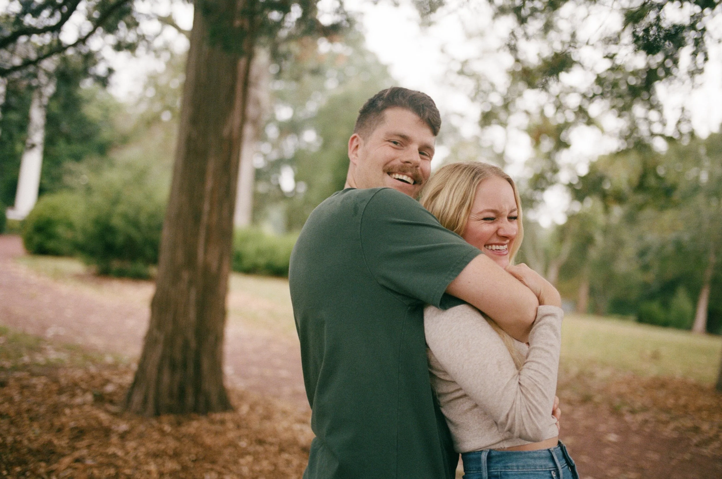 A happy couple hugging outdoors in a park, with trees and a dirt path in the background.
